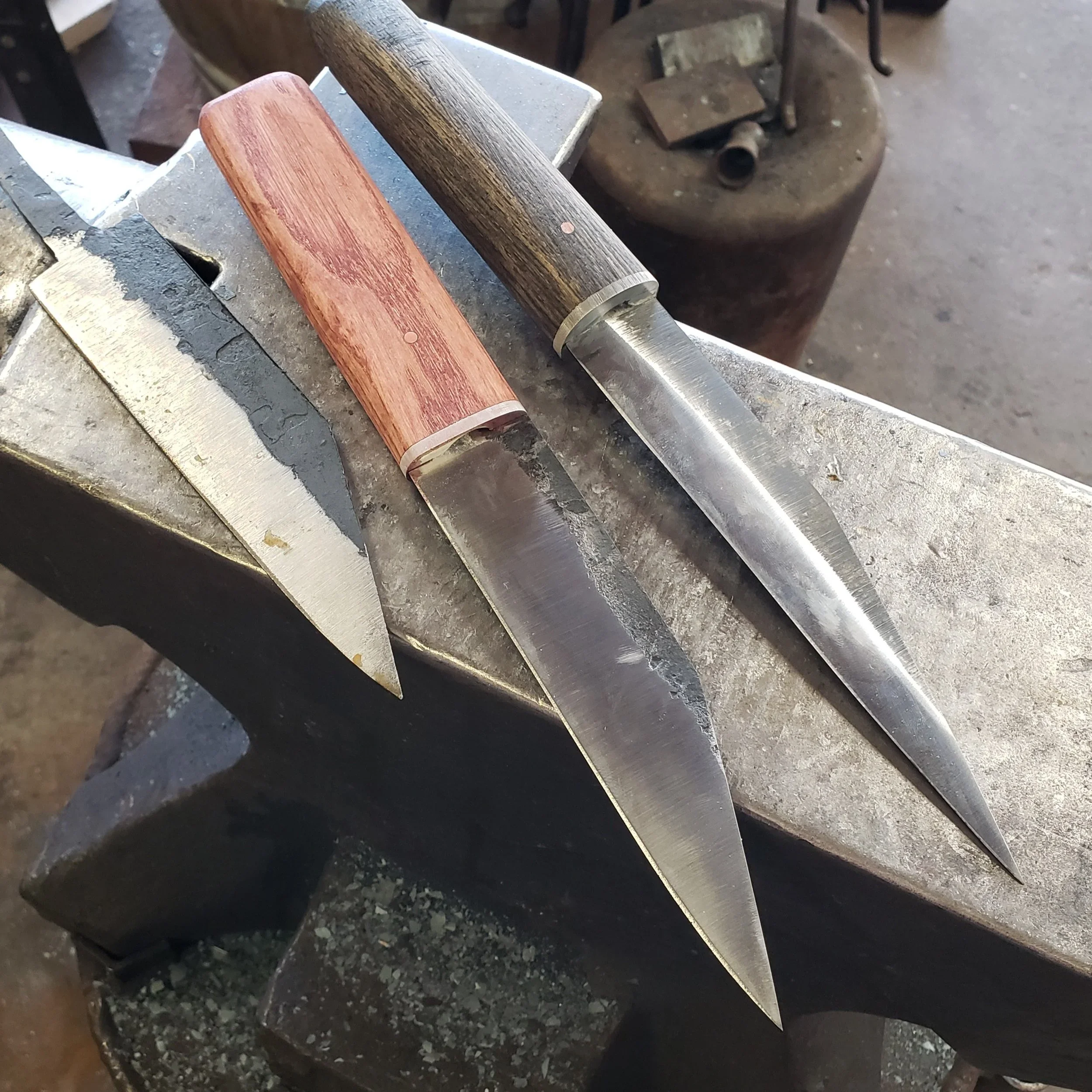 A woodworking workshop with a metal work surface, holding a partially polished wooden handle and a metal tool with a wooden grip. The background shows a workbench with a grinding wheel and various metal parts.