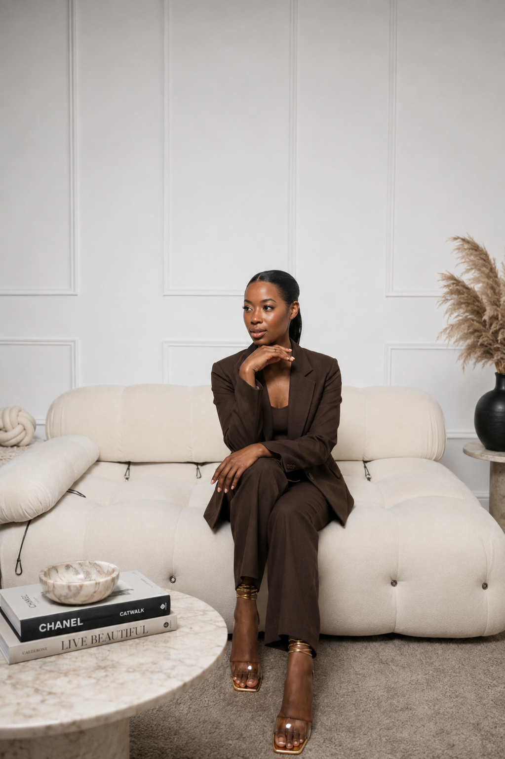 A woman in a brown suit sitting on a cream-colored couch in a modern minimalist room with white walls, a marble coffee table with books titled 'Chanel', 'Catwalk', and 'Live Beautiful', and a black vase with pampas grass.