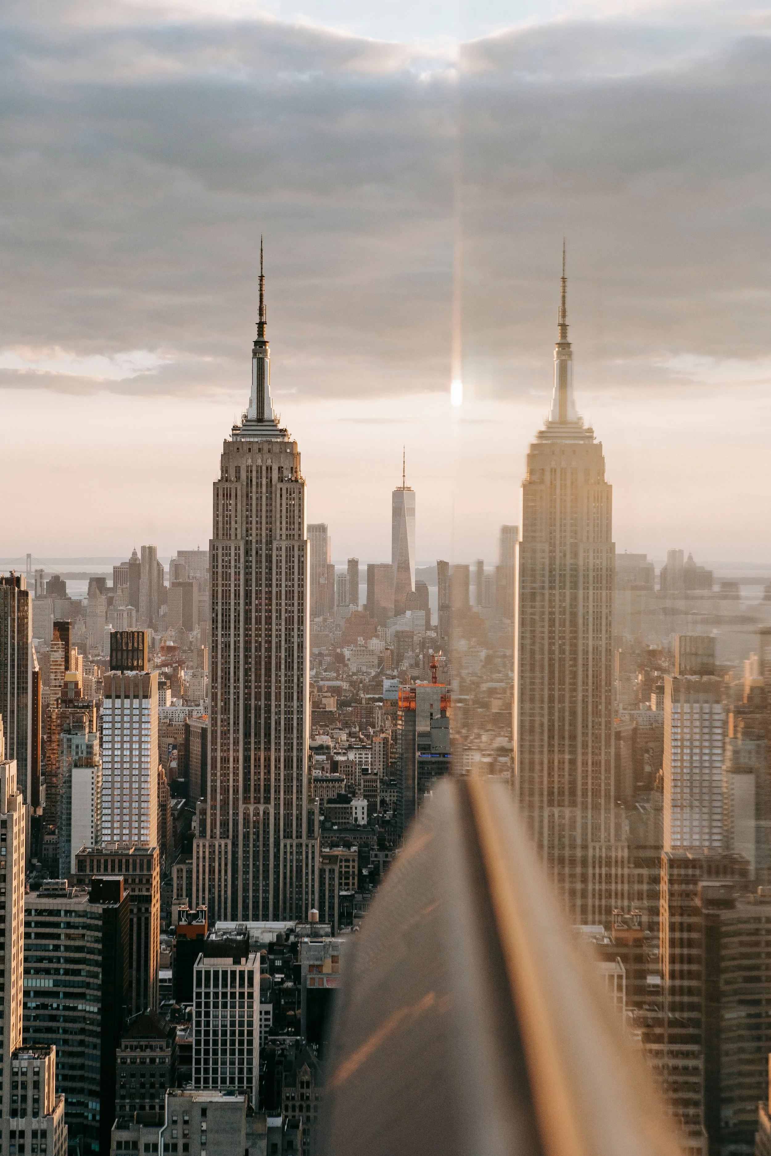 New York City skyline at sunset featuring the Empire State Building, One World Trade Center, and a mirrored reflection.