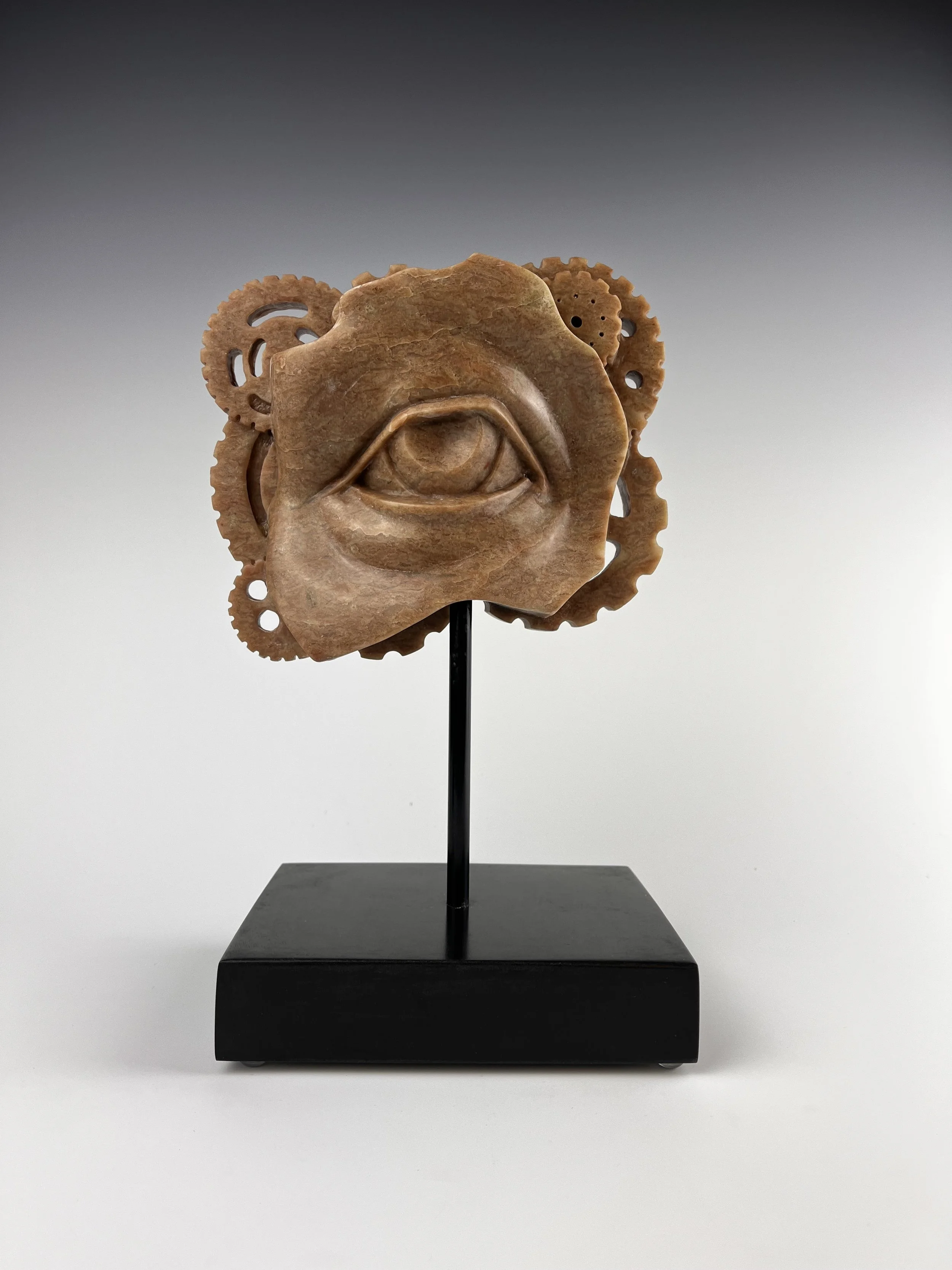 carved alabaster eye with carved gears ivisable on the edges of the sculpture, all on a neutral grey background