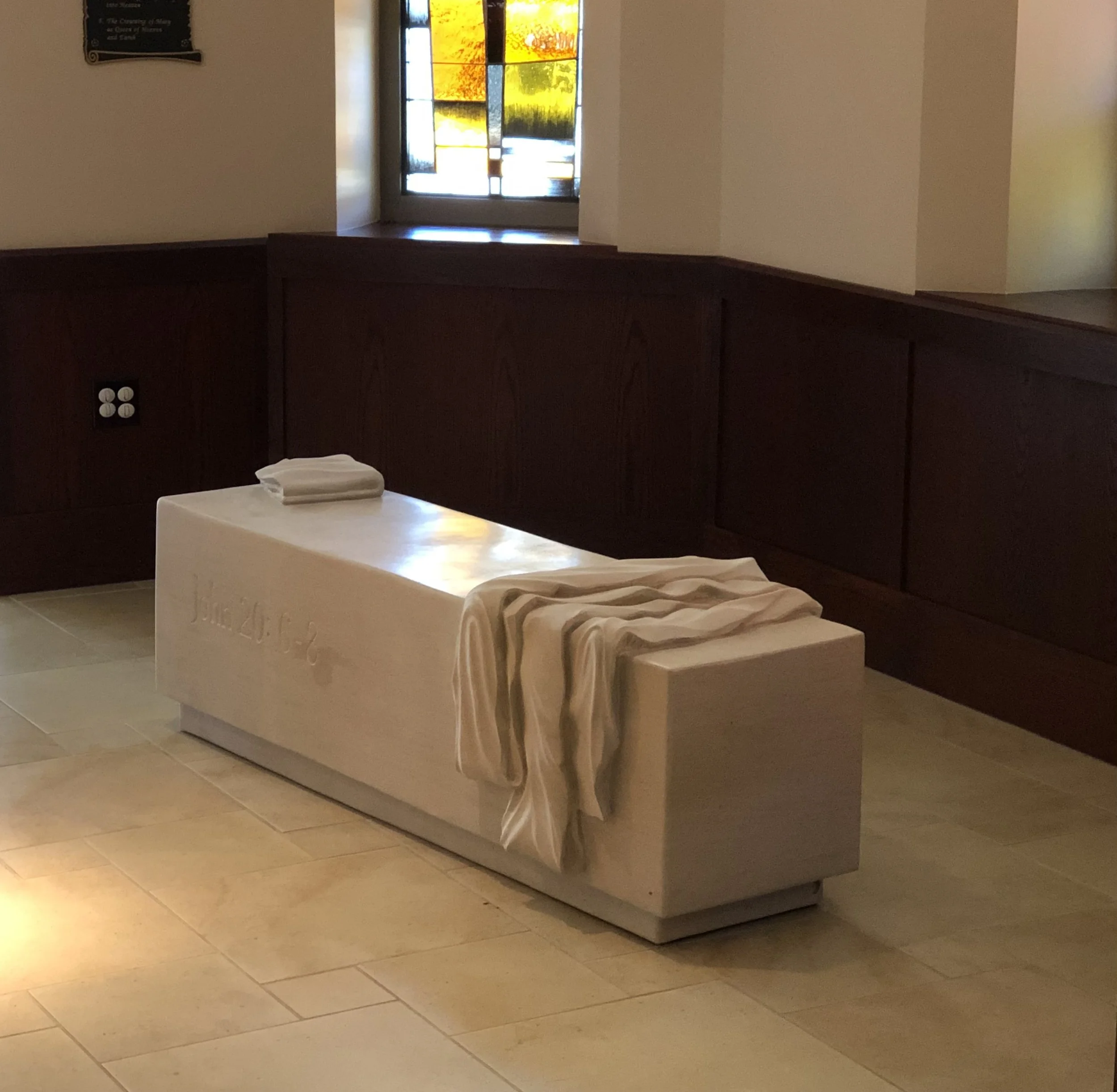 This is a Limestone sculpture with stone carved to look like the burial wrappings and face covering of Christ that serves as a bench in a Catholic Church