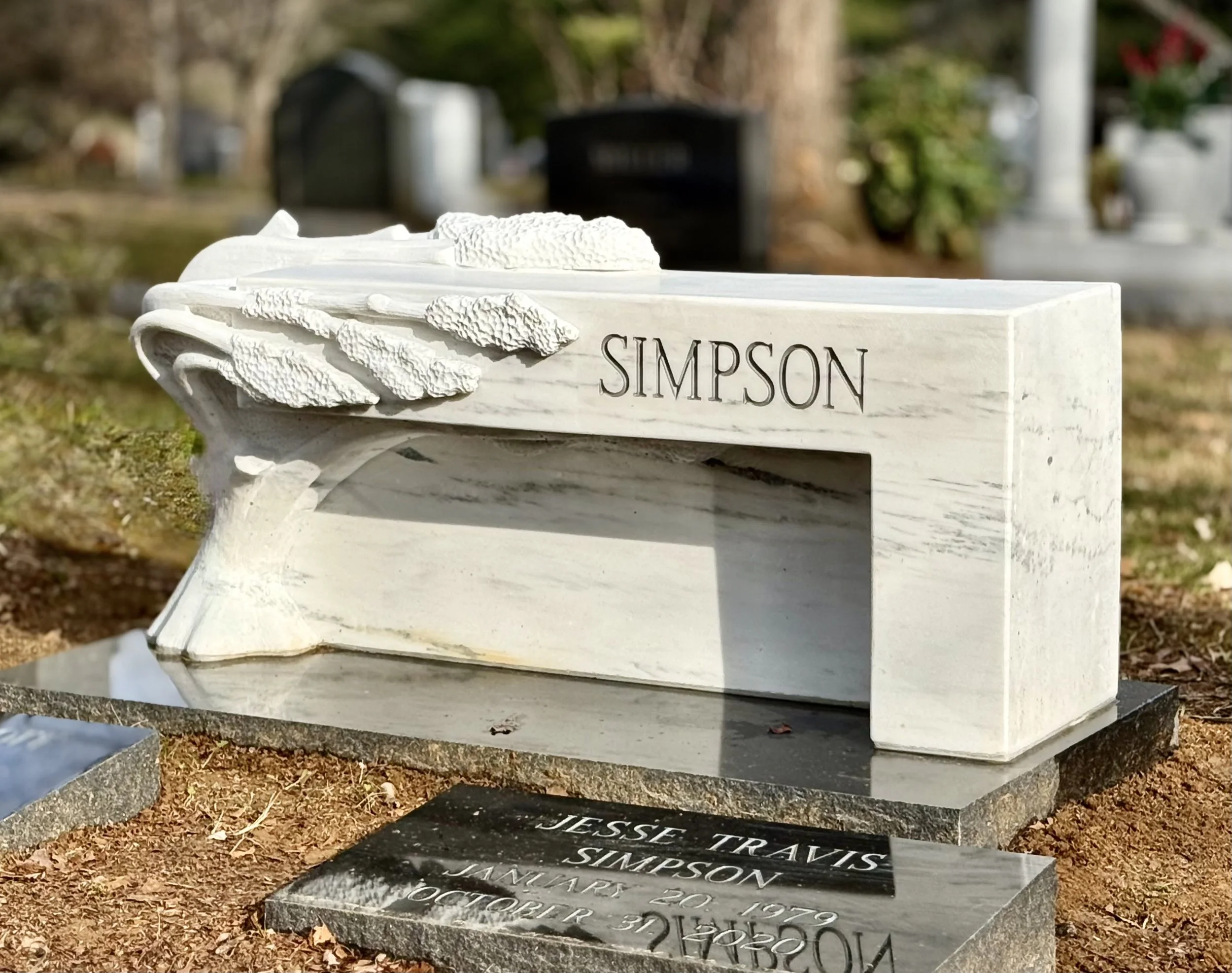 This is a sculpture that serves as a grave marker monument. It is a white marble bench with a tree emerging from it. It is sitting in a cemctery