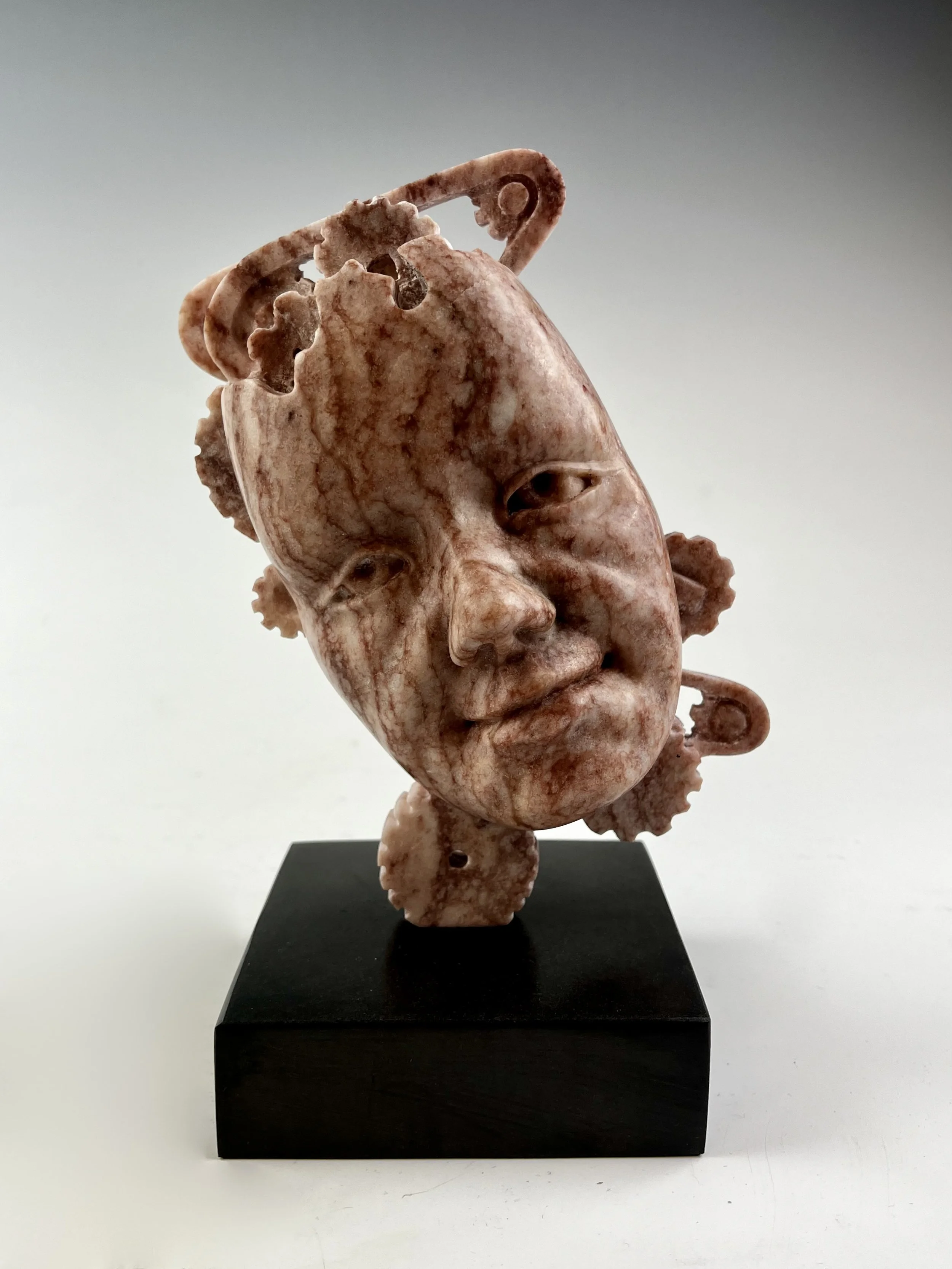 Mechanically Balanced Mind is a Raspberry Alabaster sculpture of a face with gears and belts carved in the back.