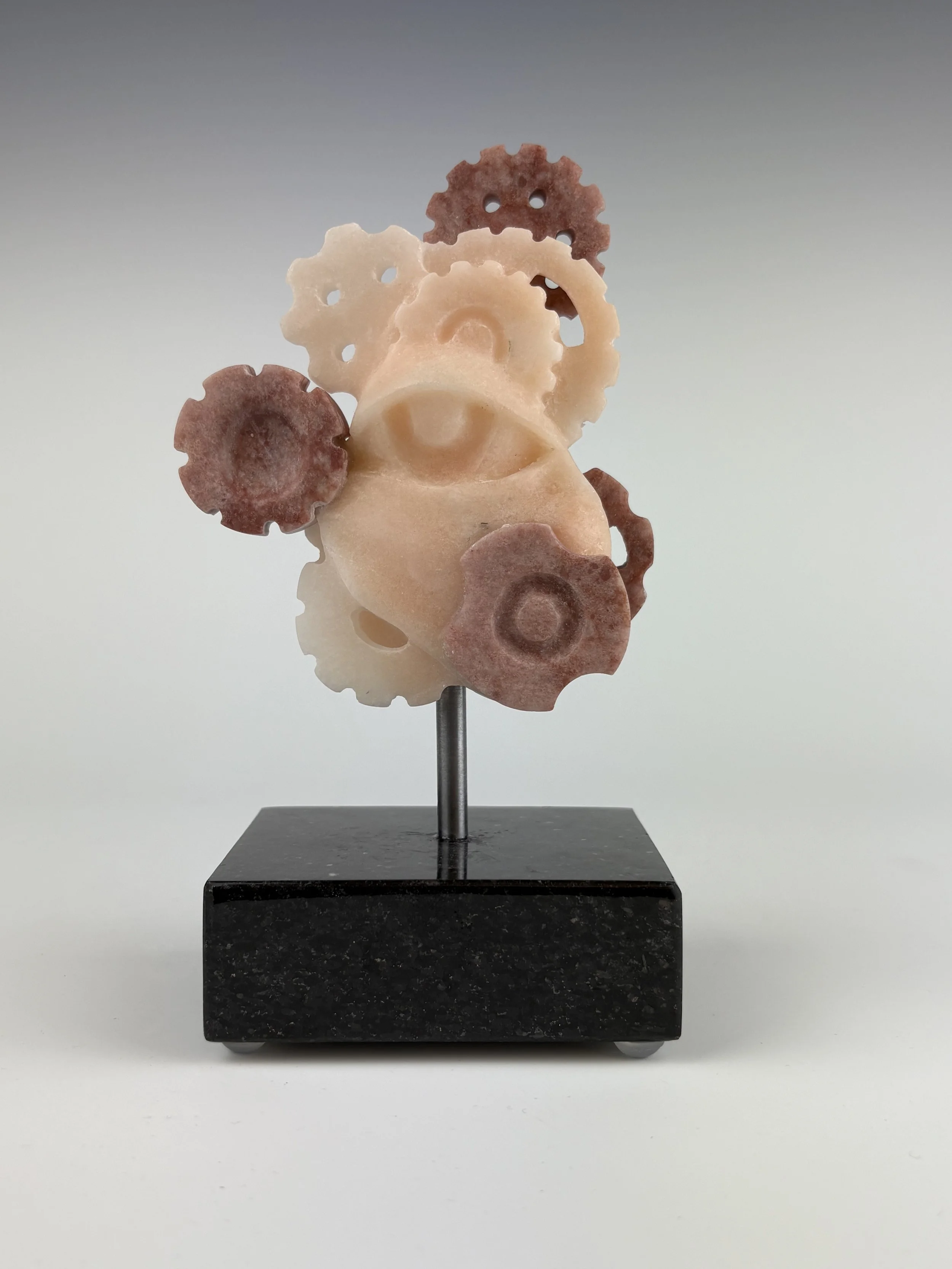 Translucent pink alabaster carved eye and gears with raspberry alabaster carved gears added to the sculpture on a steel post and stone base against a neutral background.