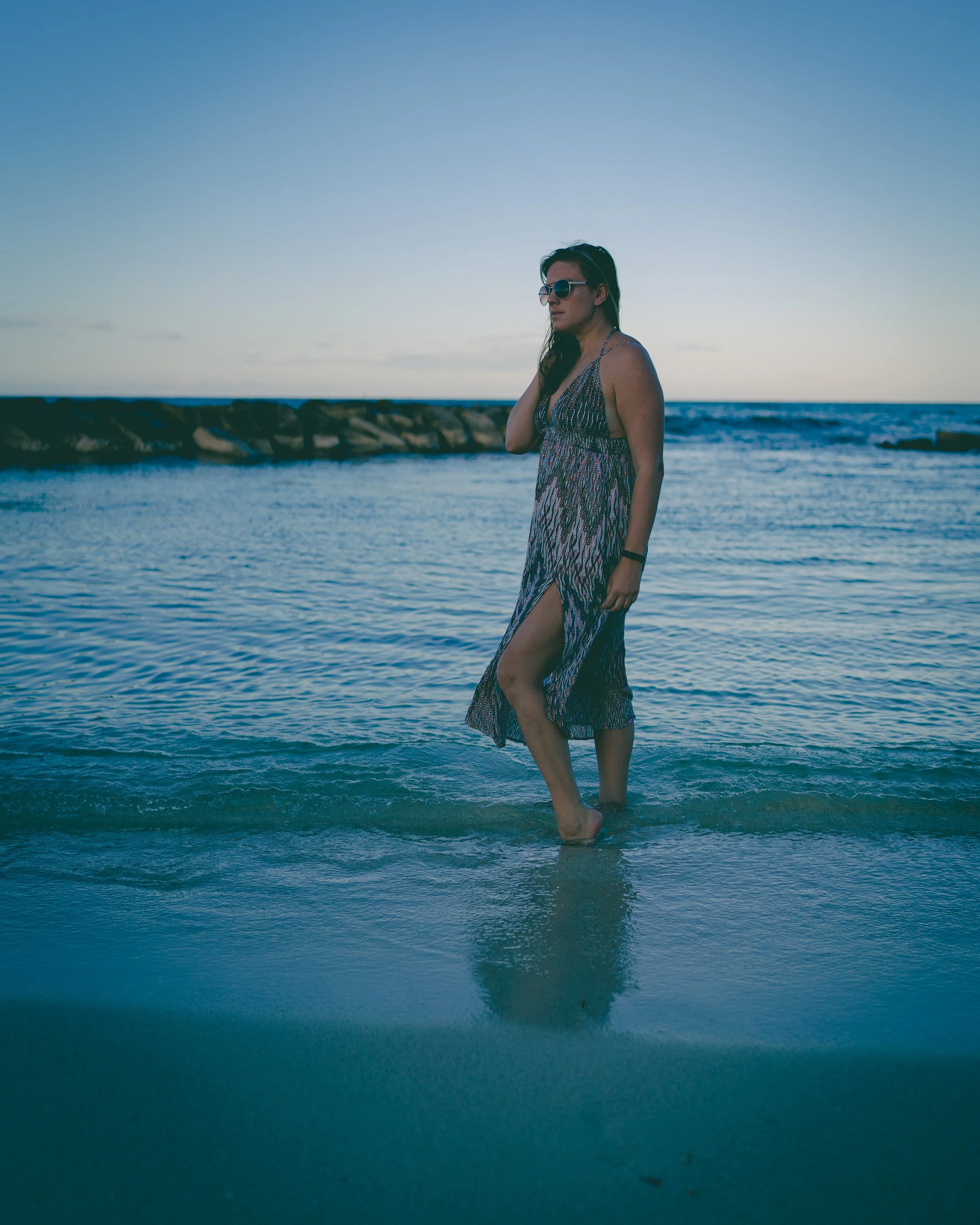 A woman with long dark hair, wearing sunglasses and a sleeveless patterned summer dress, stands in shallow ocean water near a rocky shoreline during sunset.