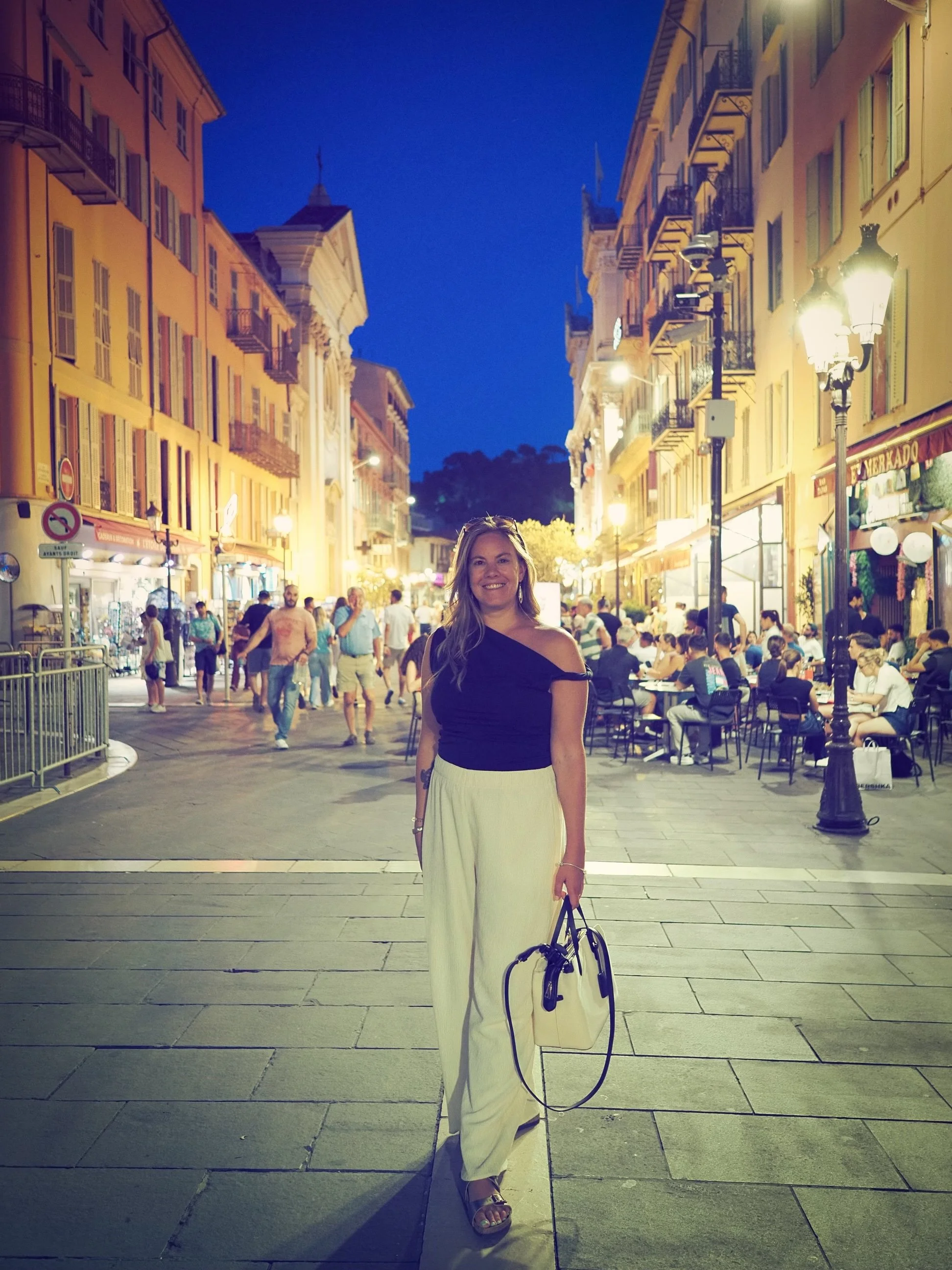A woman smiling and standing on a busy street at night, surrounded by restaurants, shops, and people seated at outdoor tables, with historic buildings and street lamps illuminating the scene.