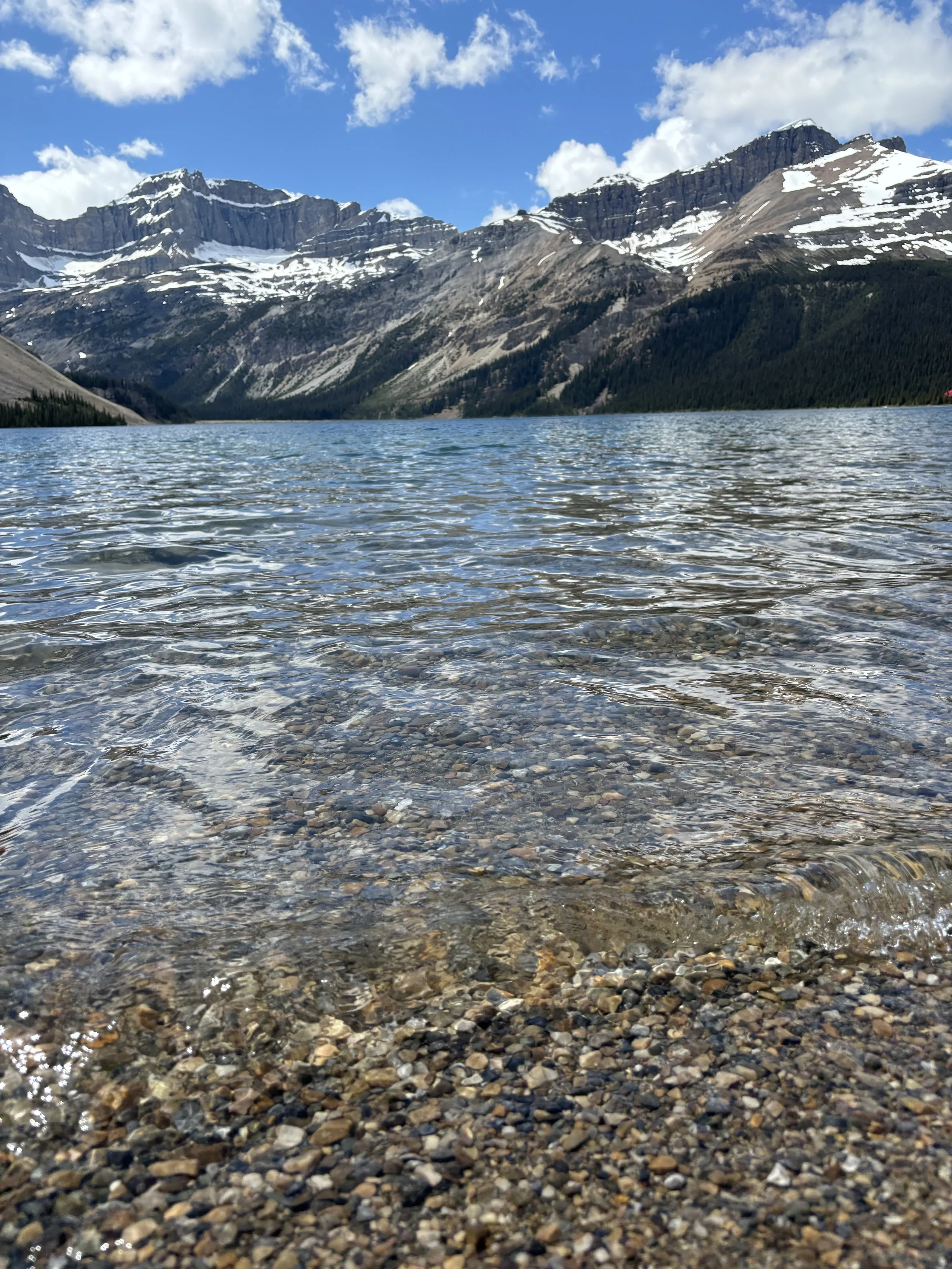 A clear lake with pebbles near the shore, surrounded by mountains with snow patches under a partly cloudy sky.