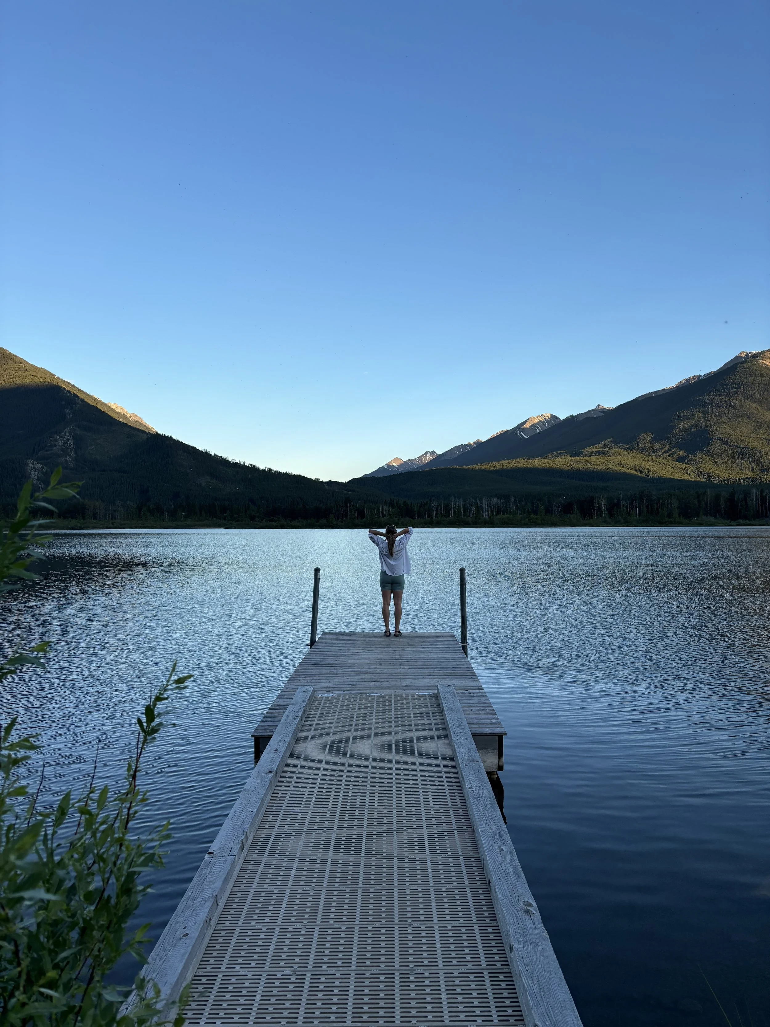 A person standing at the end of a wooden dock on a calm lake, with mountains and a clear blue sky in the background.