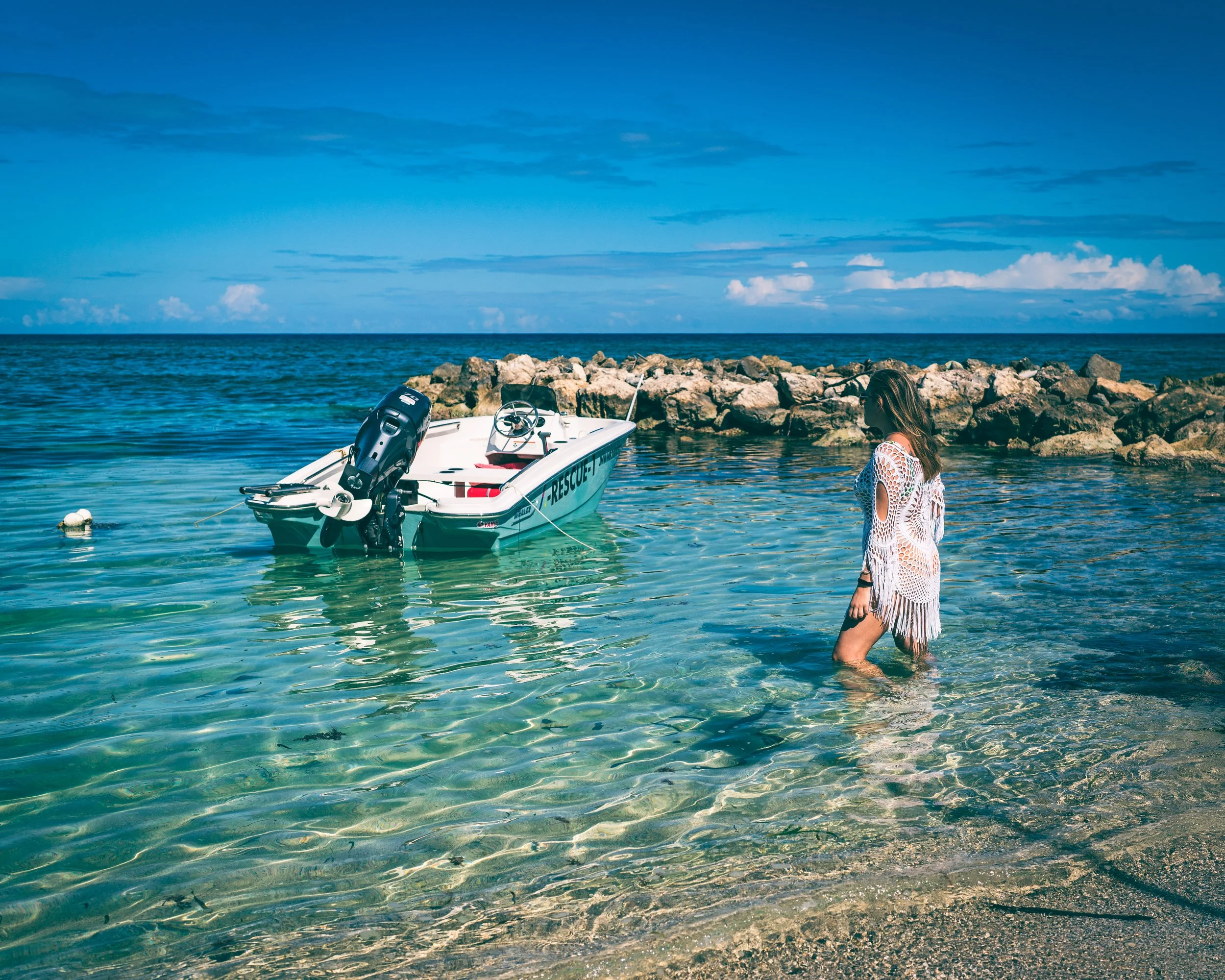 A woman in a white crochet dress stands in shallow clear water near a rocky shoreline, with a small motorboat anchored nearby, against a blue sky with scattered clouds.