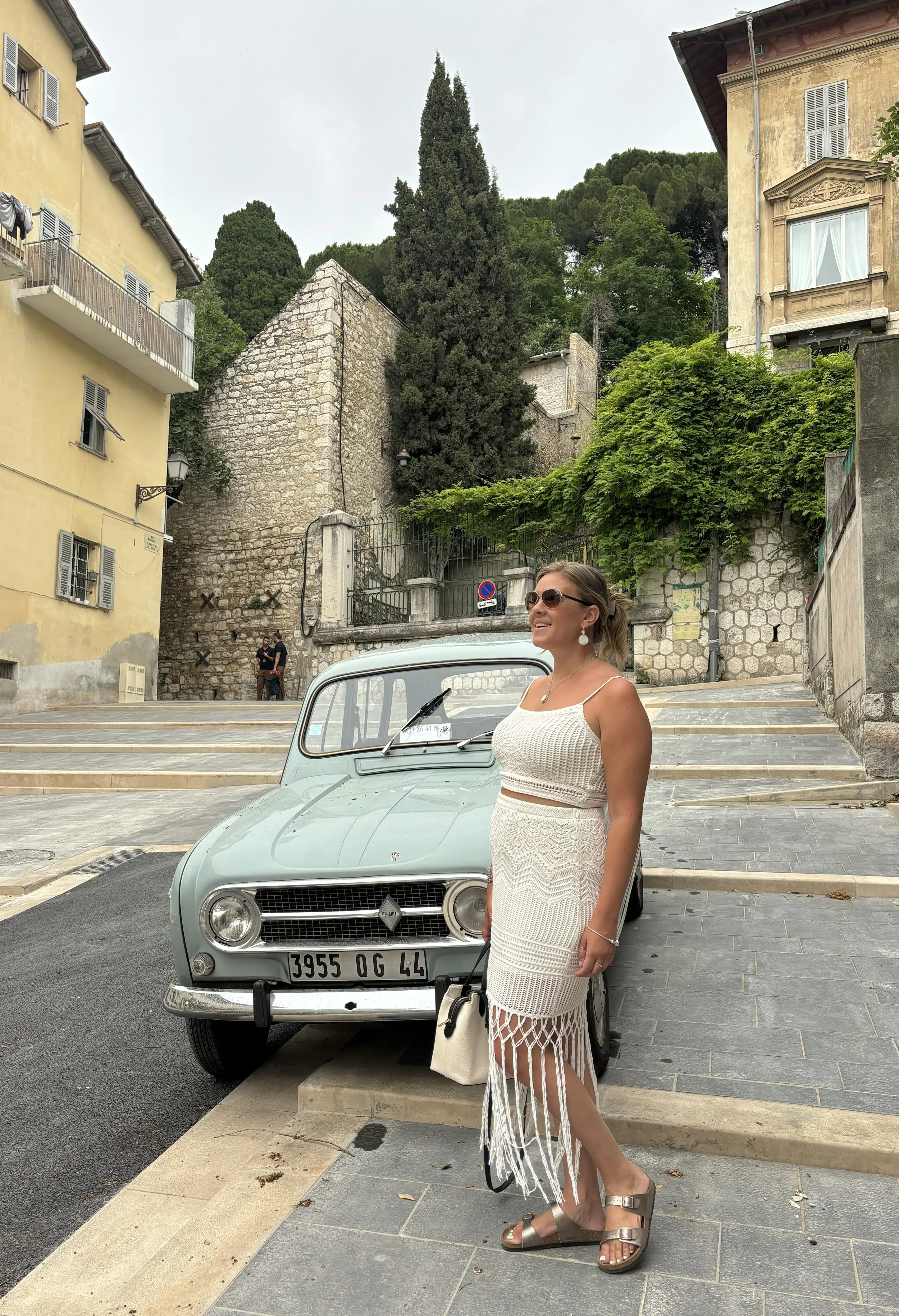 A woman in a white crocheted dress and sunglasses is standing next to a vintage light green Renault car with French license plates, in an outdoor European city street with historic buildings, greenery, and a stone staircase in the background.
