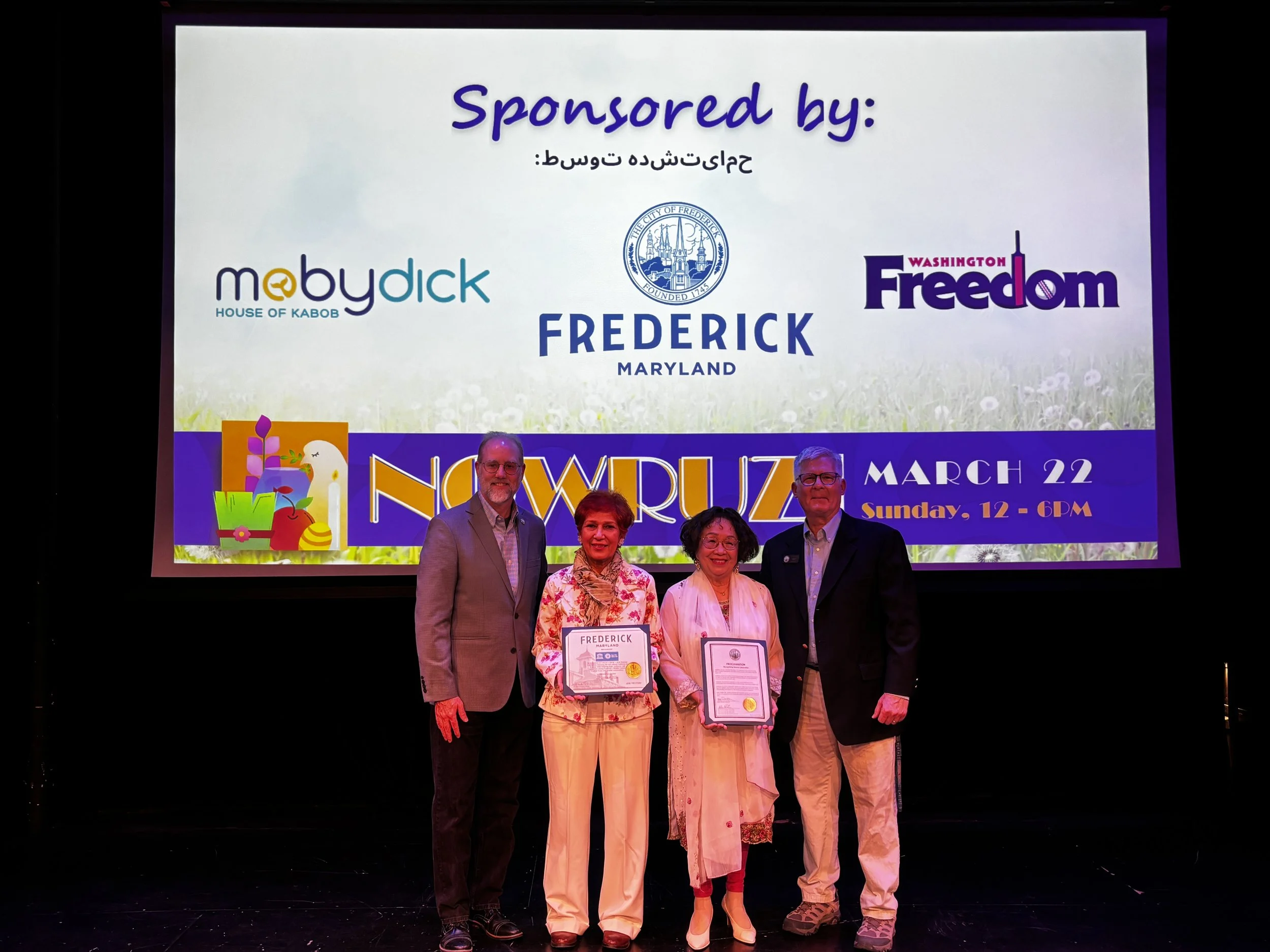 four people posing for picture holding two certificates in front of screen that reads: "sponsored by" accompanied by company logos