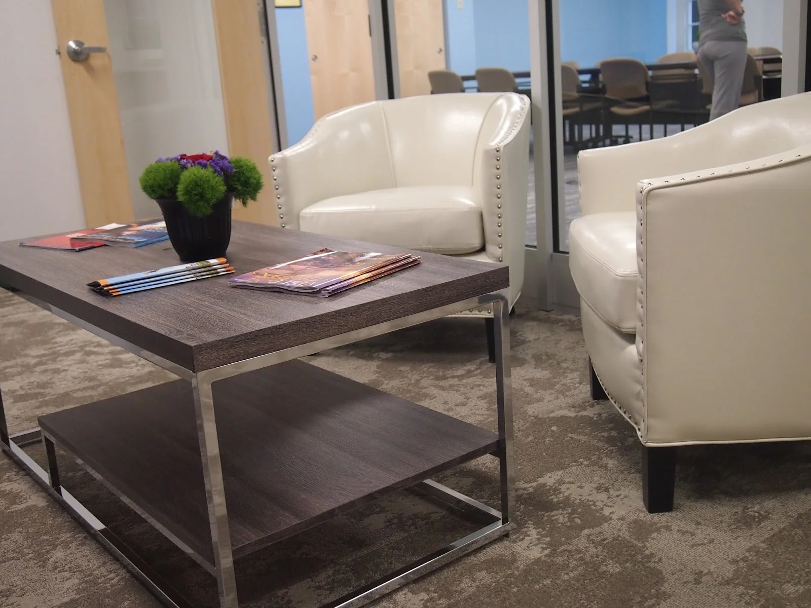 Stylish modern seating area with two white leather armchairs, a sleek wooden coffee table holding magazines, and a vase of vibrant greenery and flowers.