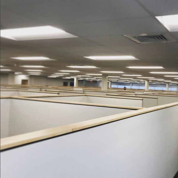 A vast office space filled with uniform, empty cubicles under bright fluorescent lights. One person is partially visible in the distance, creating a sense of isolation.