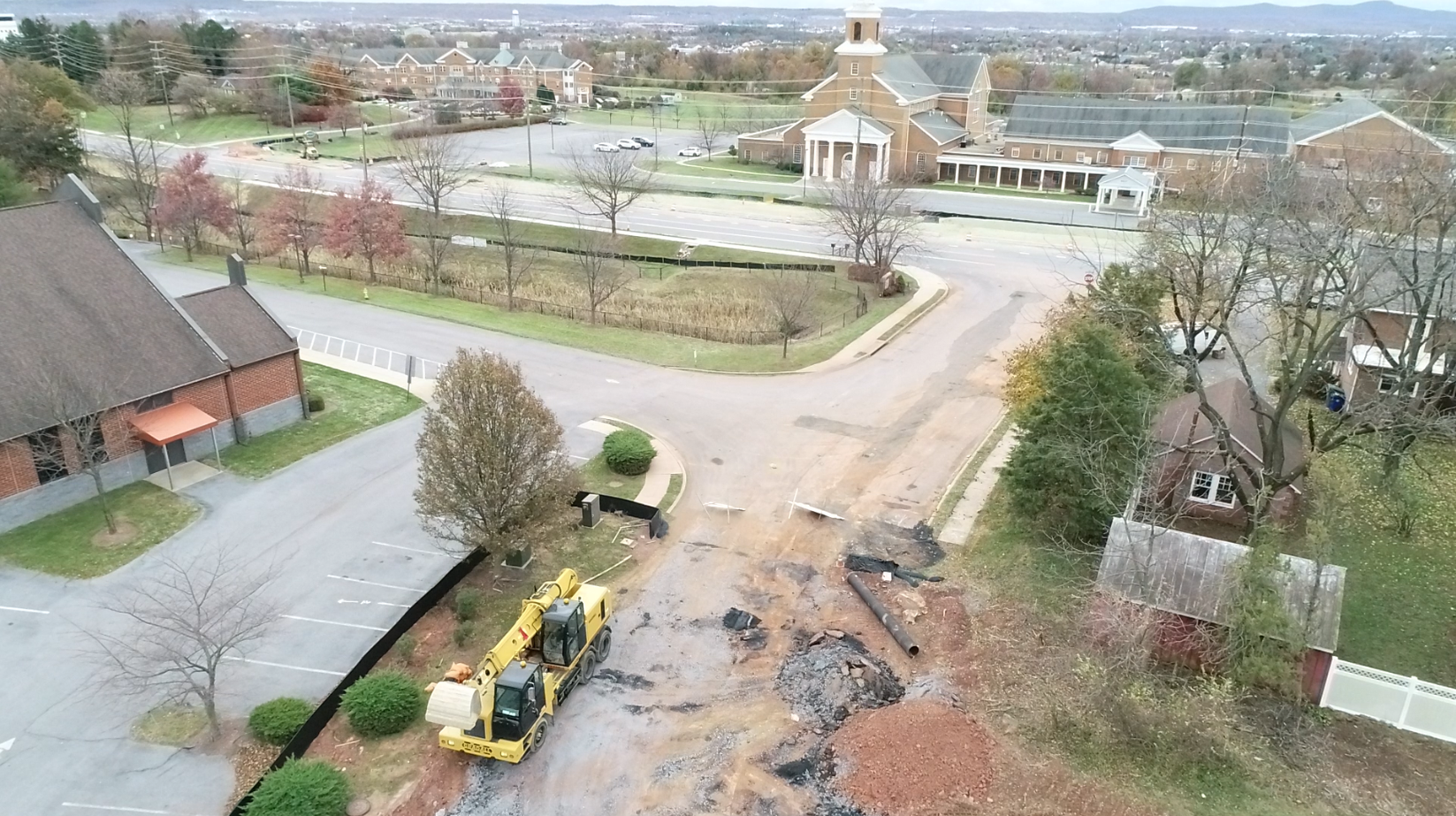Aerial view of construction with a yellow excavator and damaged road in front of a brick building with a church in the background..png