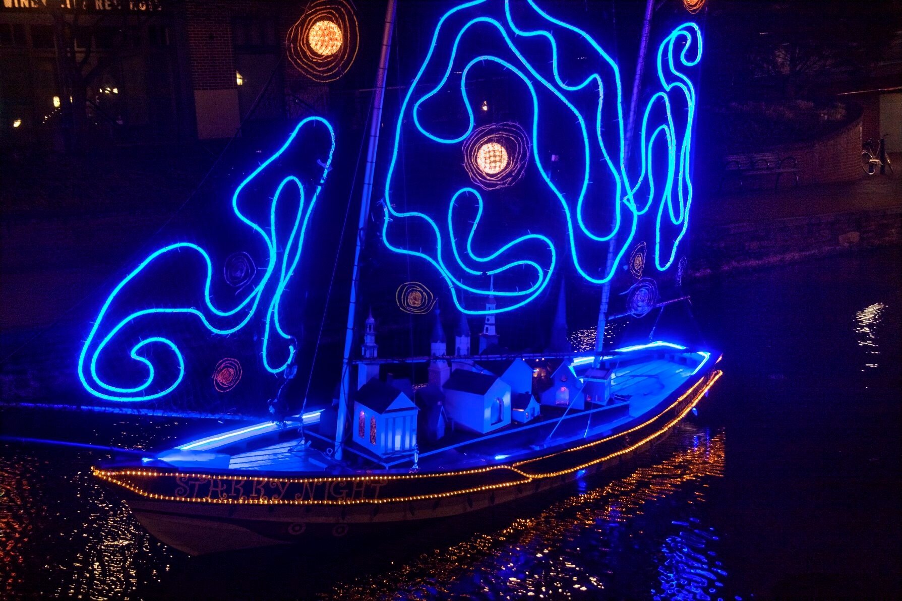 A decorated sailboat illuminated with bright blue neon lights, featuring illuminated models of buildings on its deck and reflecting in dark water at night.