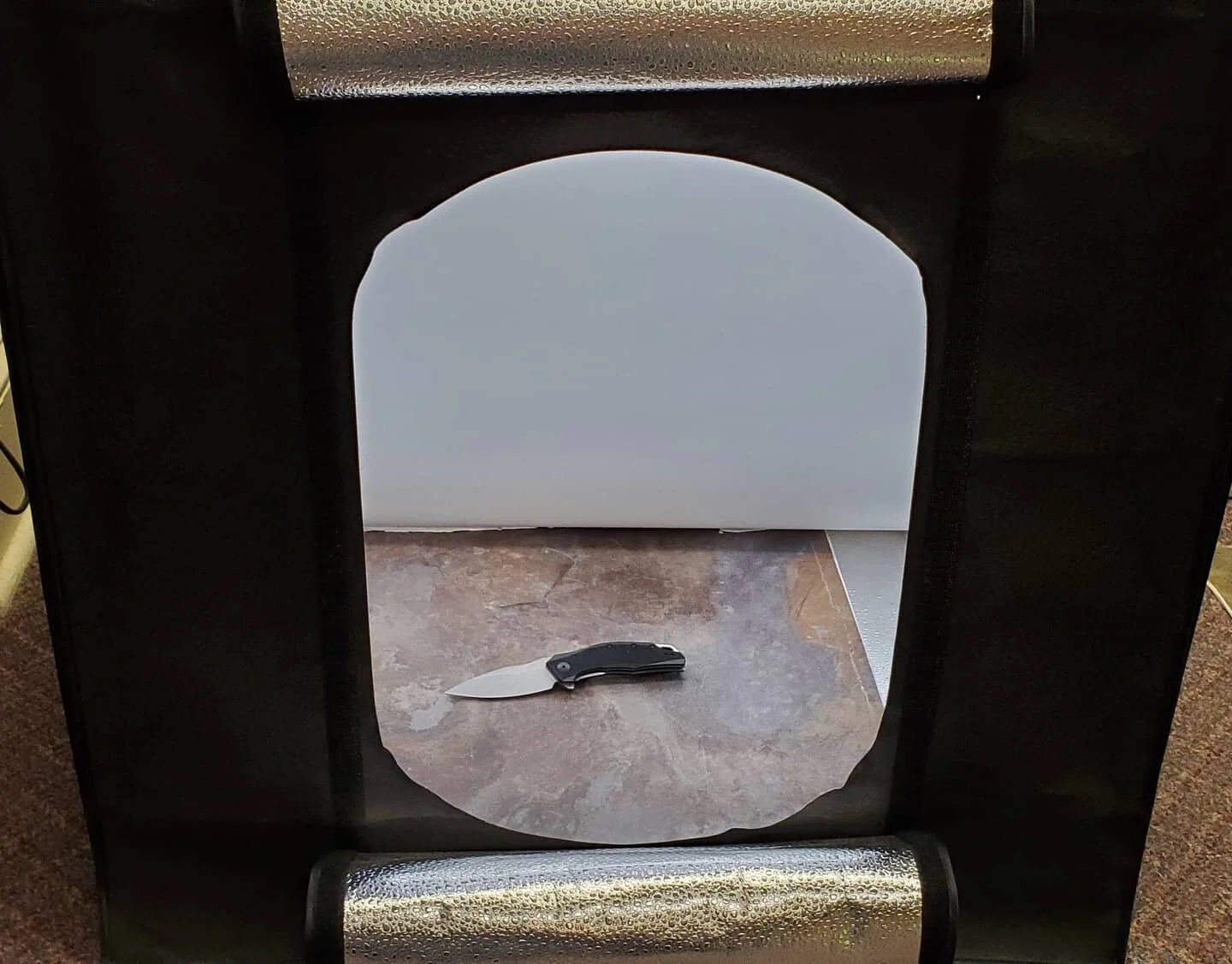 A black folding pocket knife lies closed on a textured brown and gray surface, viewed through an opening in a black photography light tent with silver reflective interiors.