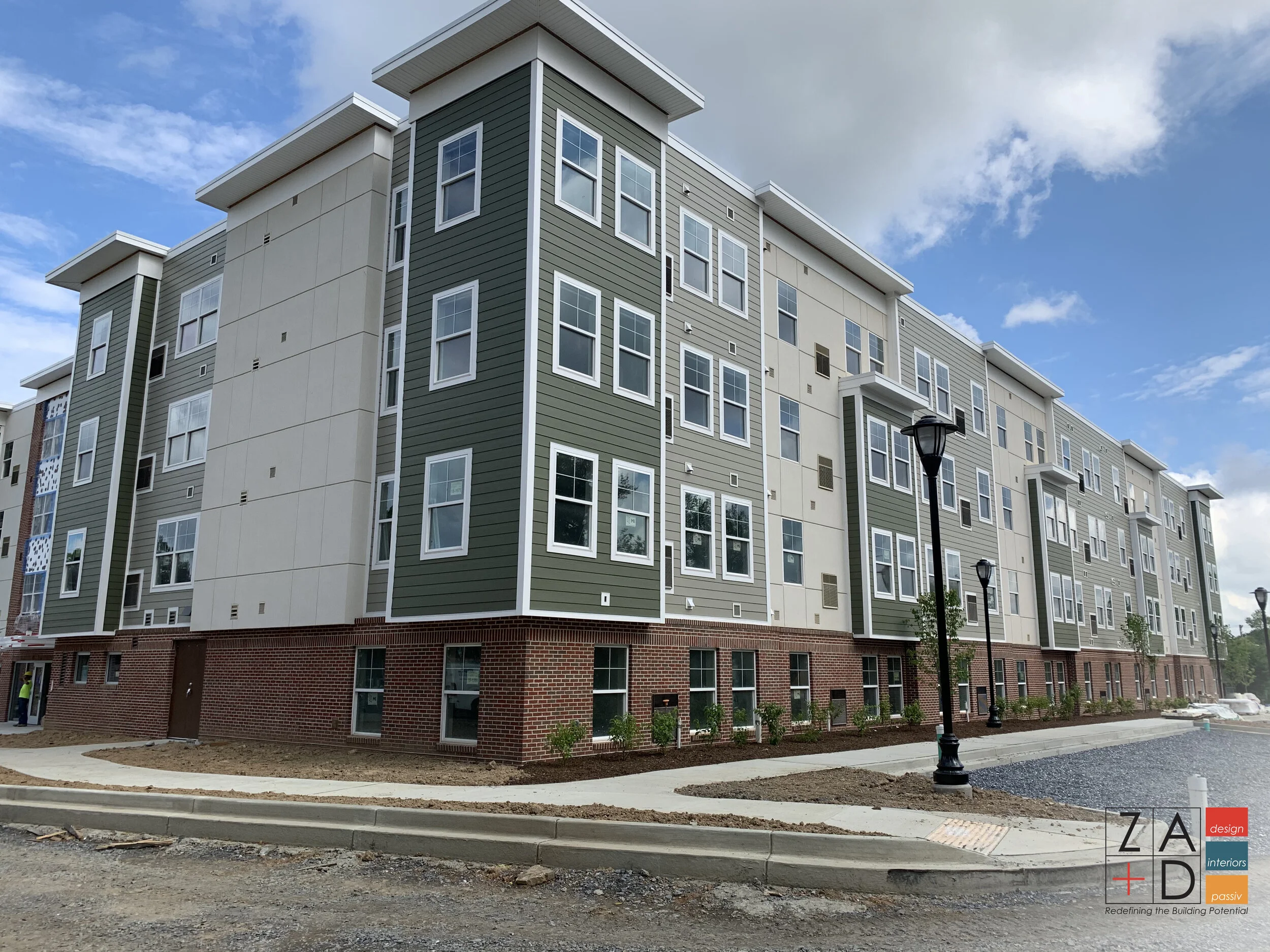 A modern, multi-story apartment building with a light grey and dark green siding exterior, brick base, and numerous windows under a partly cloudy blue sky..JPG
