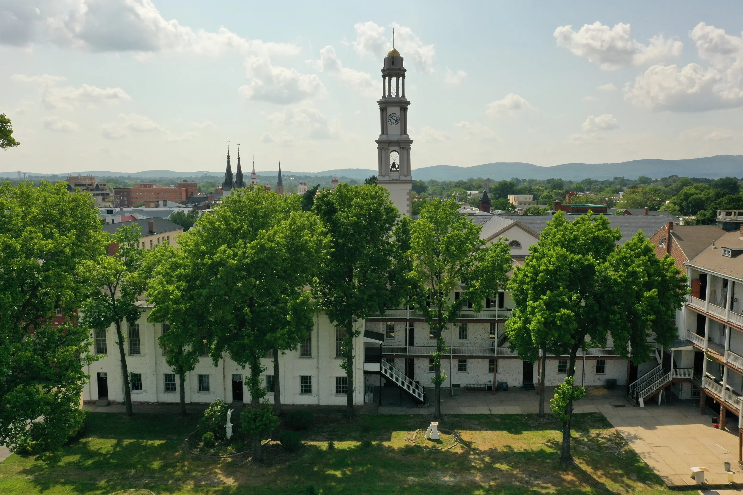 Aerial view of historic white buildings with multiple floors and balconies, surrounded by lush green trees, with a tall clock tower and church spires visible in the background under a cloudy sky..jpg