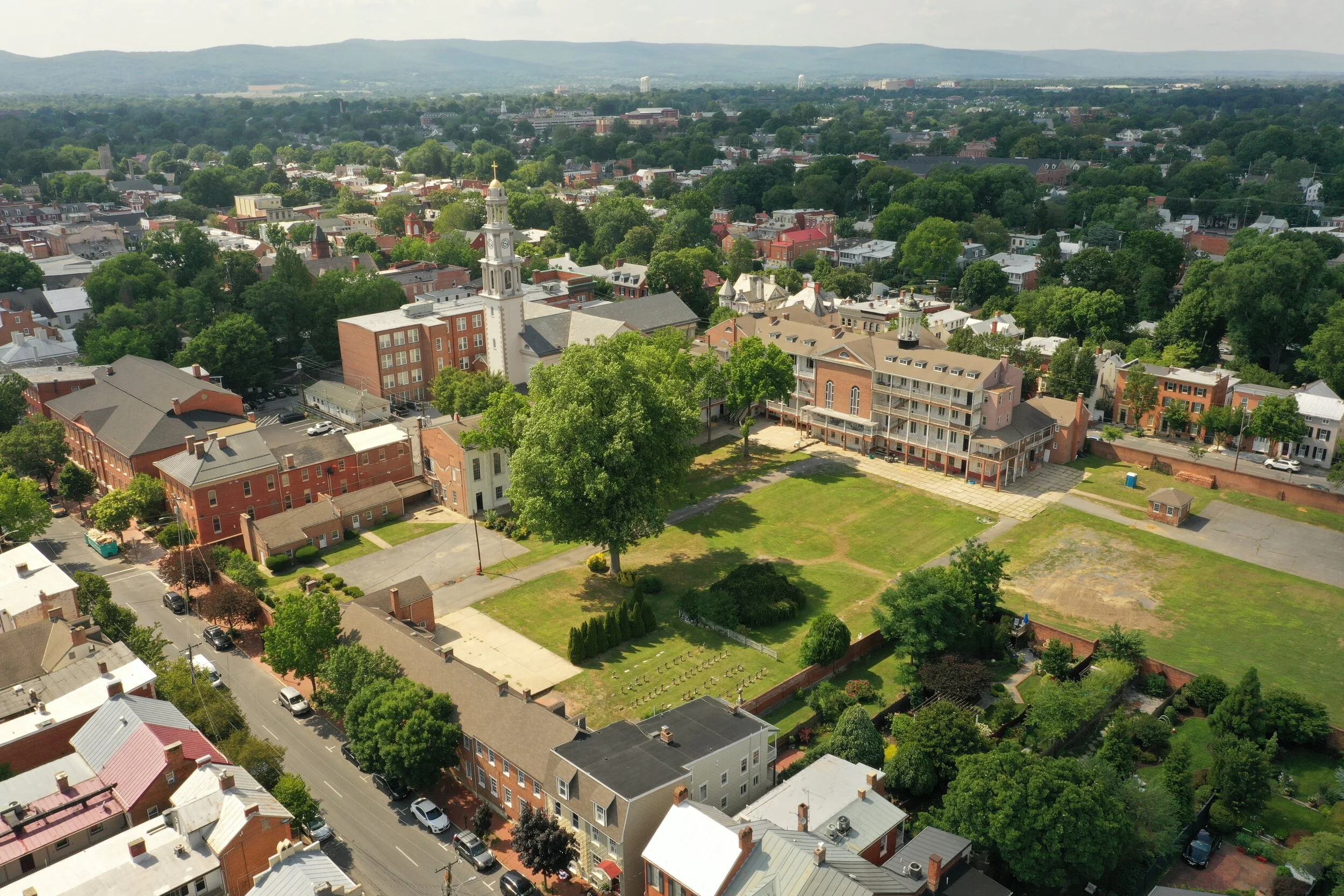 Aerial view of a historic town square with a white church steeple, brick buildings, a large grassy field, and surrounding trees..jpg