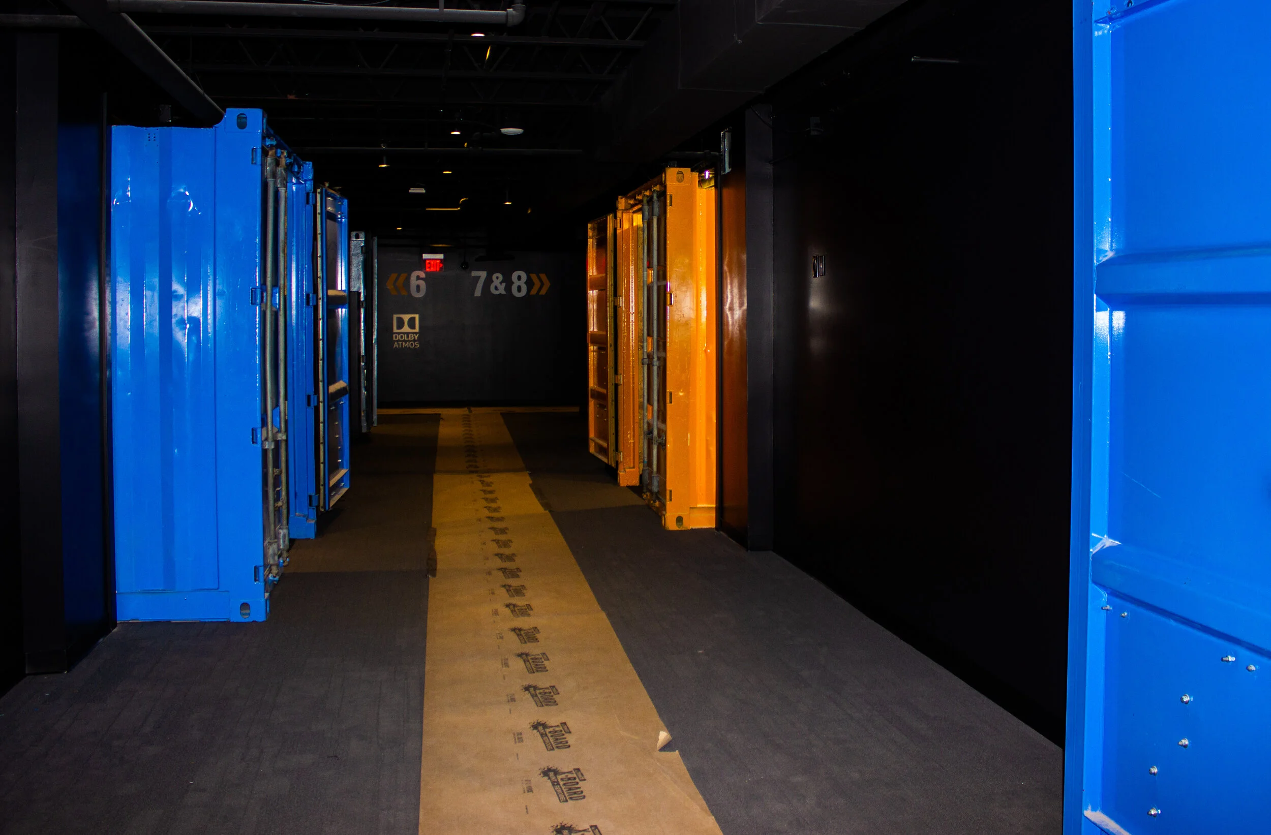 A dimly lit hallway with bright blue and orange shipping containers lining the walls, resembling private movie theaters or rooms..jpg