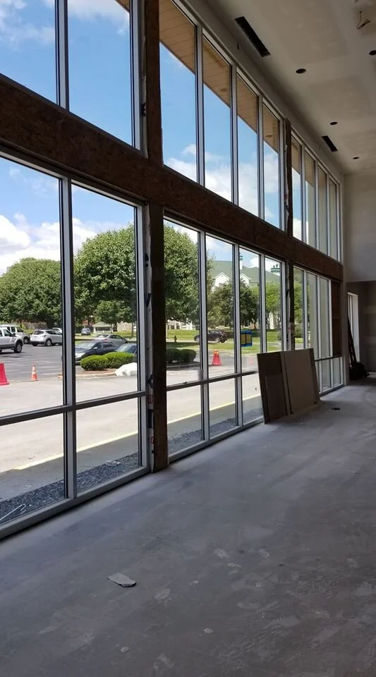Interior view of a large commercial building under construction, featuring a wall of floor-to-ceiling windows that reflect a bright blue sky with scattered clouds. Through the windows, a parking lot with several cars, green trees, and buildings are visibl.jpg