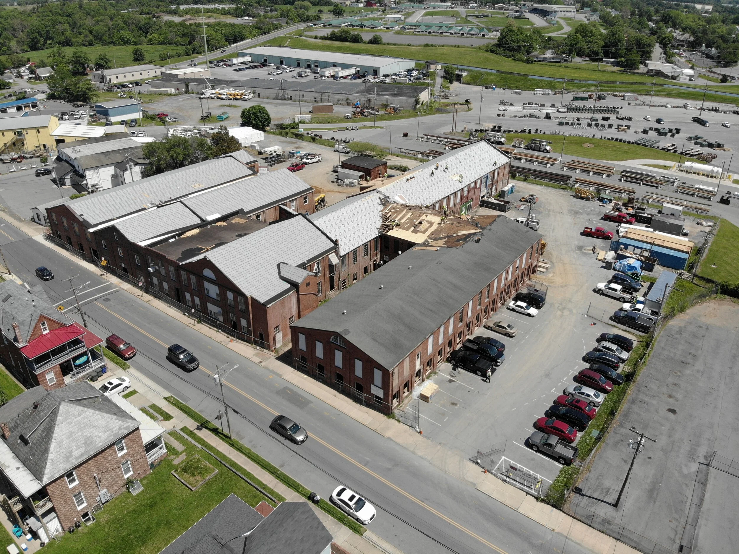 Aerial view of a complex of brick buildings with gray metal roofs, showing some roof damage on one building, surrounded by asphalt parking lots with cars and adjacent residential homes and industrial yards..jpg