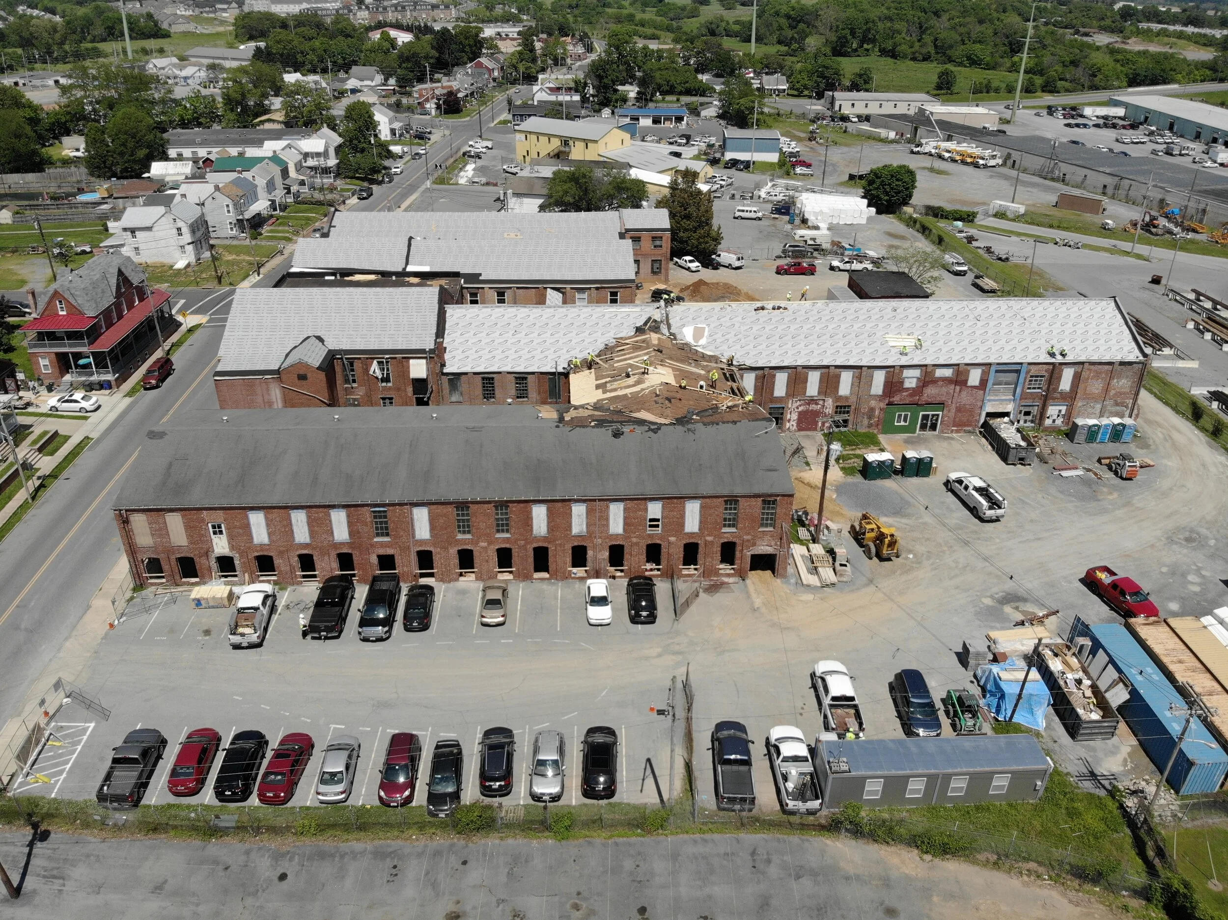 Aerial view of a complex of brick buildings with gray metal roofs, showing some roof damage on one building, surrounded by asphalt parking lots with cars and adjacent residential homes and industrial yards..jpg