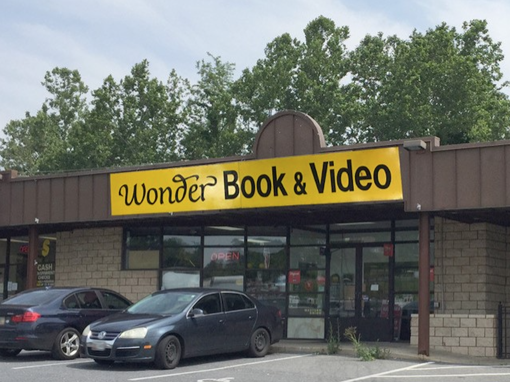 A storefront with a yellow sign reading "Wonder Book & Video" above large glass windows and doors. Two cars are parked in front of the store..png