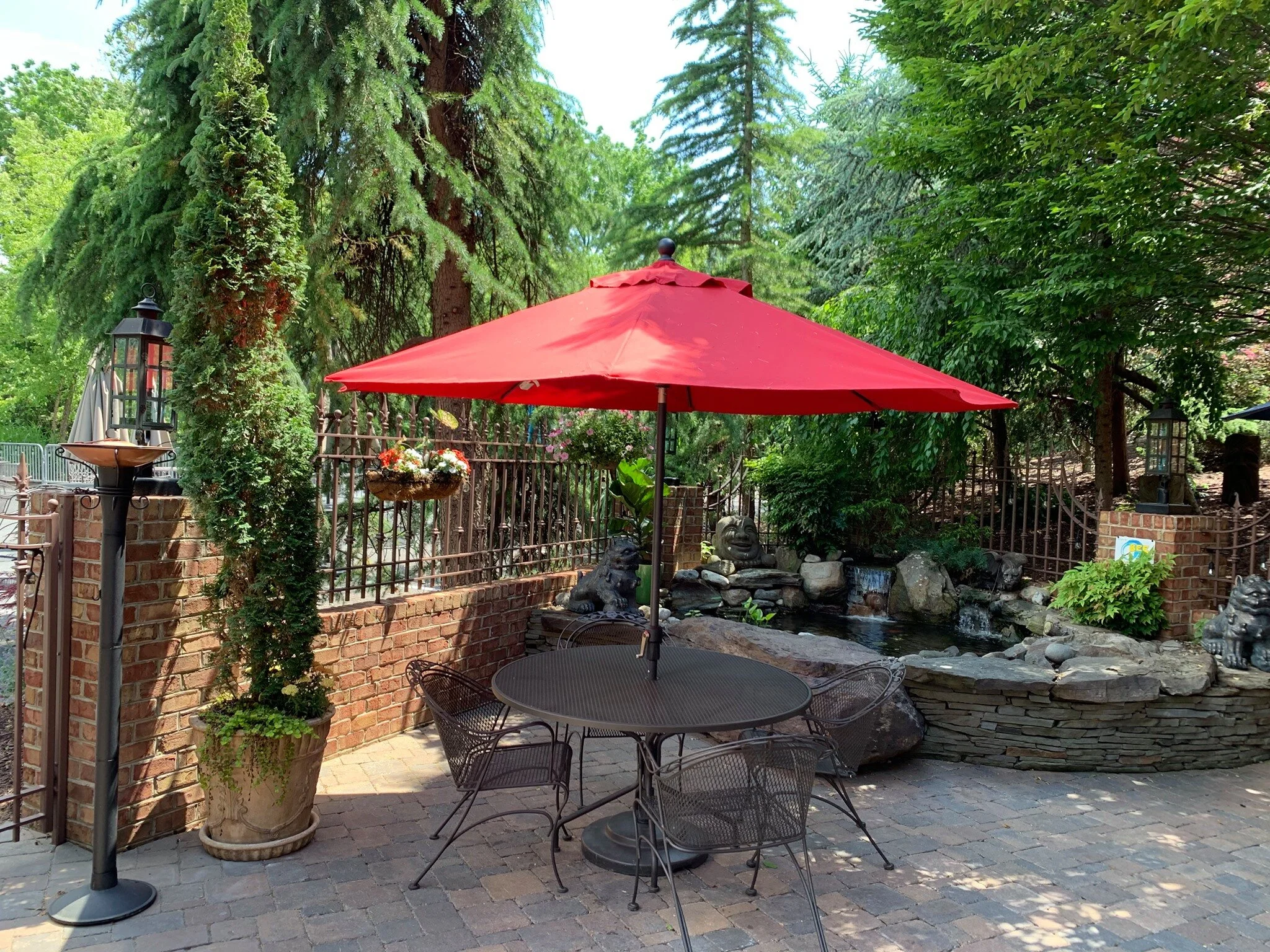 An outdoor patio with a red umbrella over a metal table and chairs, a decorative pond with a small waterfall, and lush greenery..jpg