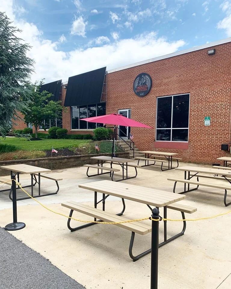 Outdoor seating area with multiple picnic tables in front of a brick building with a "Dragon Distillery" logo, featuring a pink umbrella and yellow caution ropes..jpg
