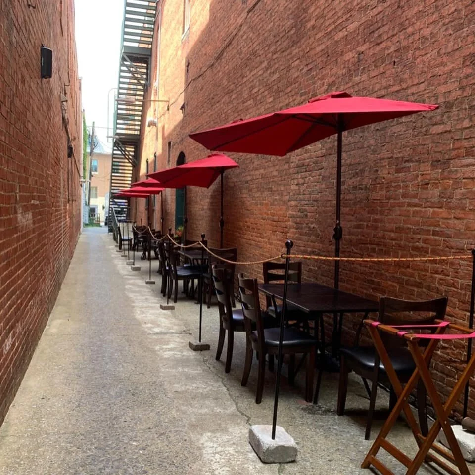 Outdoor dining setup in a narrow brick alleyway with tables and chairs under red umbrellas..jpg