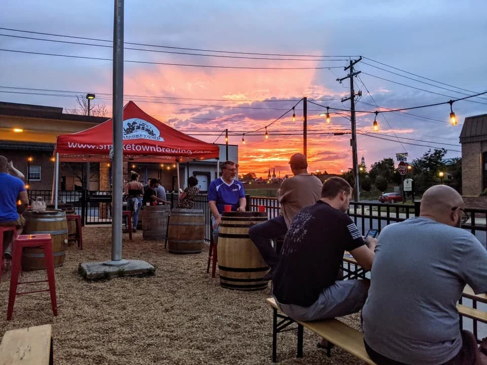 An outdoor seating area of a brewery with a red tent, barrels for tables, and people enjoying the sunset..jpg
