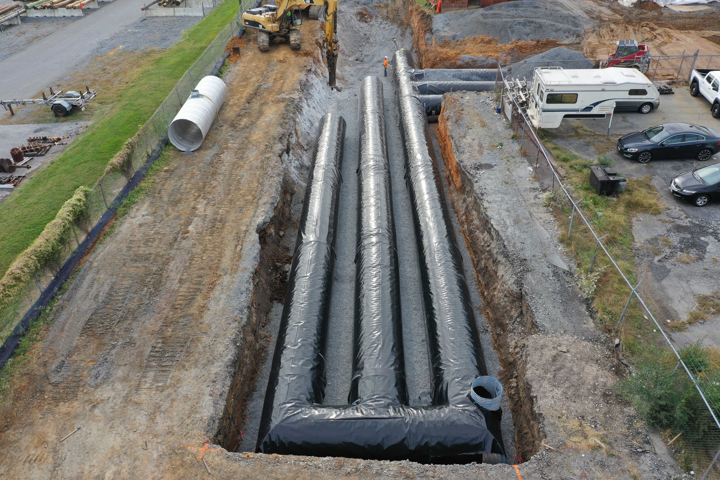 Construction site with large black pipes laid in a deep trench, surrounded by gravel. Excavator working nearby, and vehicles parked to the right..jpg