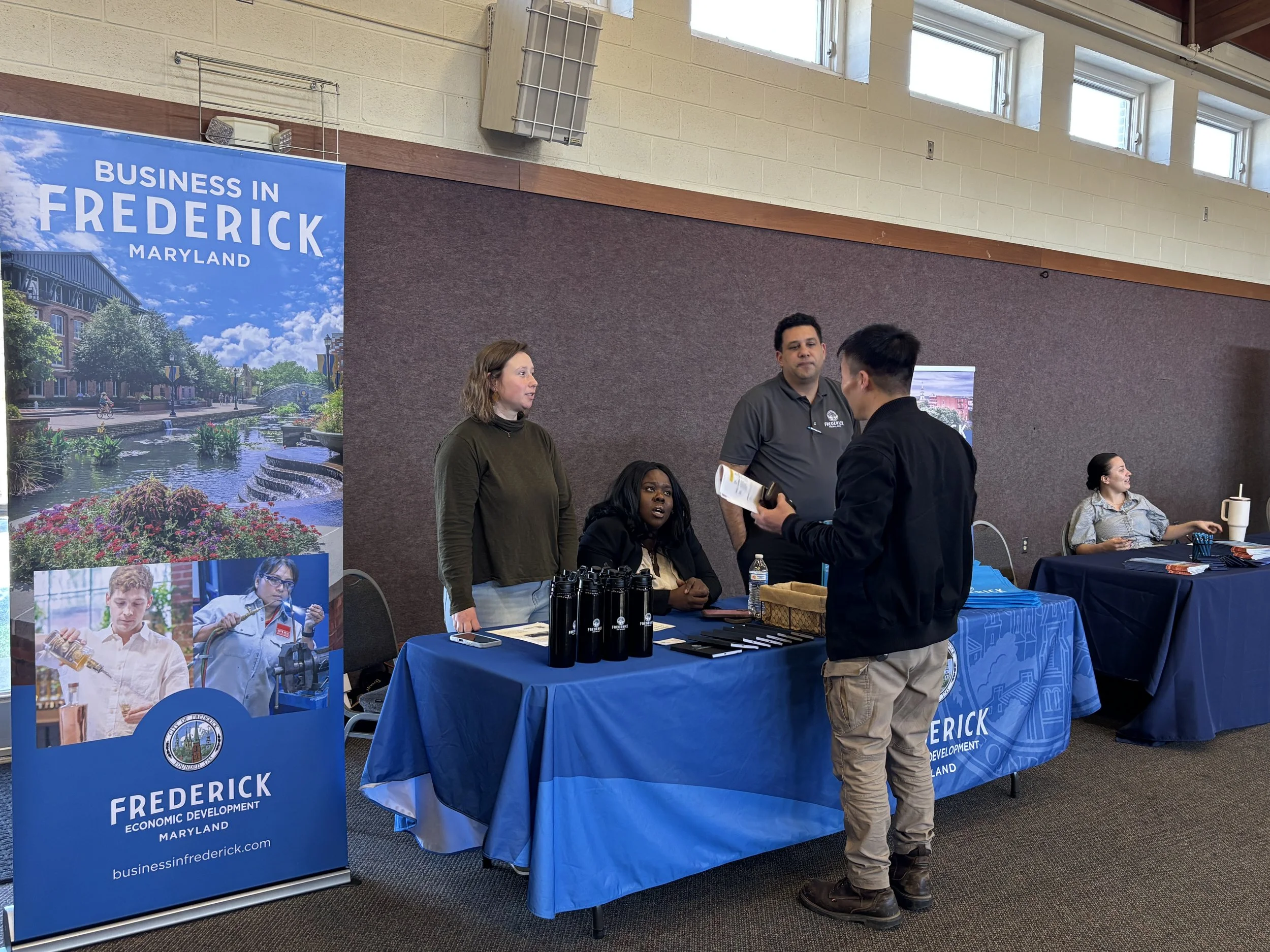 three people behind blue exhibition booth talking to one person on other side. blue Business in Frederick banner to left of table.