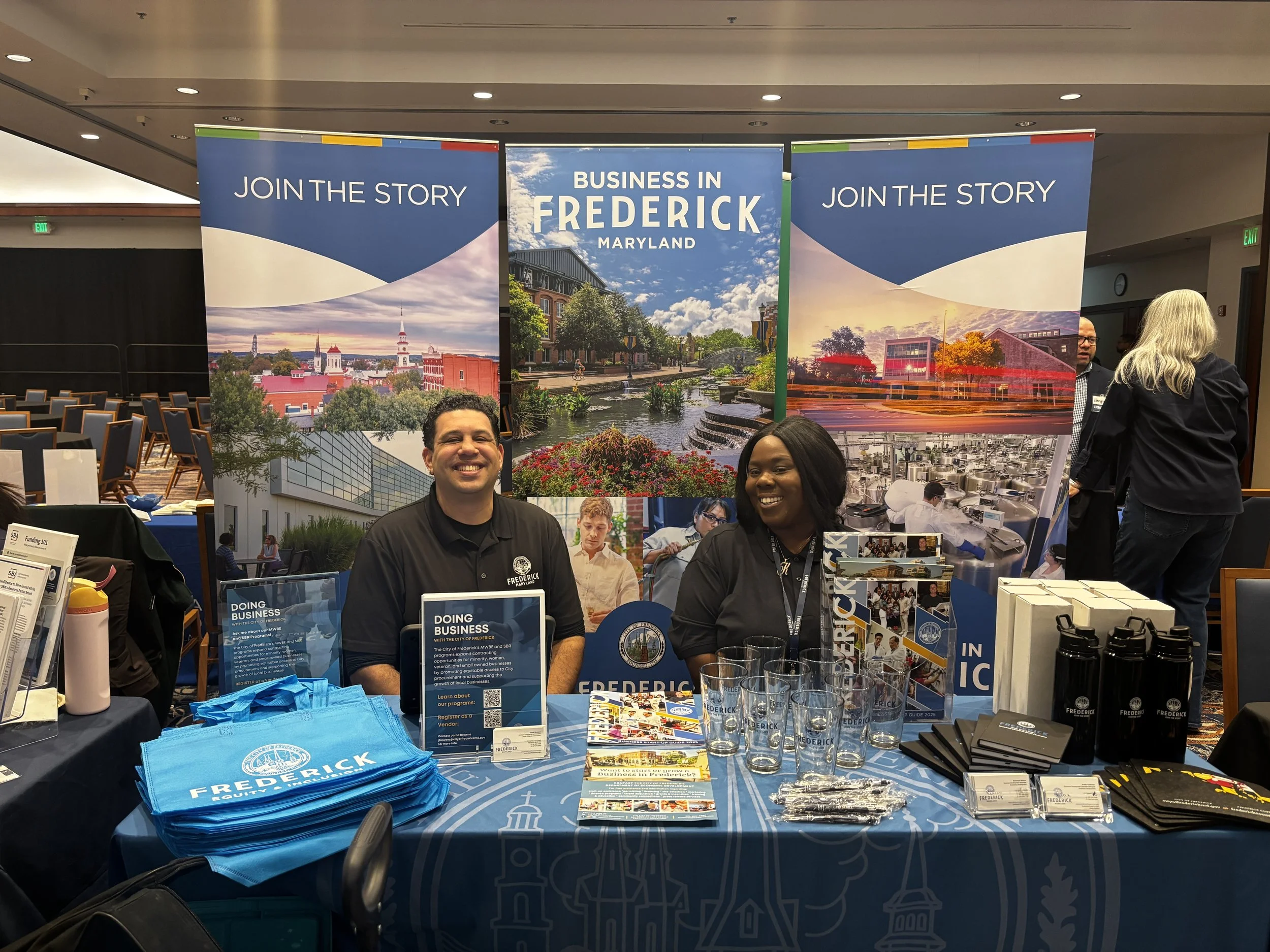 two people behind blue exhibition booth with banners that read 'join the story' and 'business in frederick maryland'