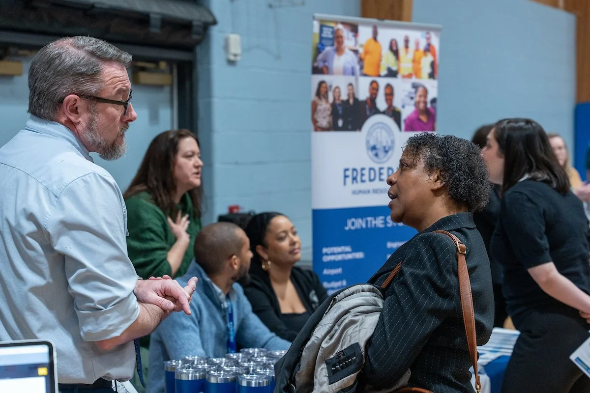 multiple people interfacing at job fair, few individuals standing and sitting behind exhibitor table talking to attendees