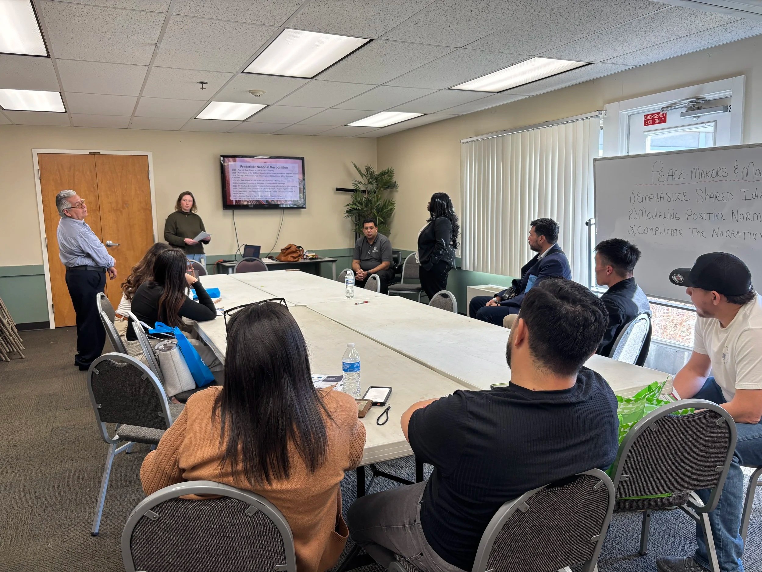 two women one man standing in front of small audience sitting around table giving presentation from tv display behind them in conference room setting, window and door on right