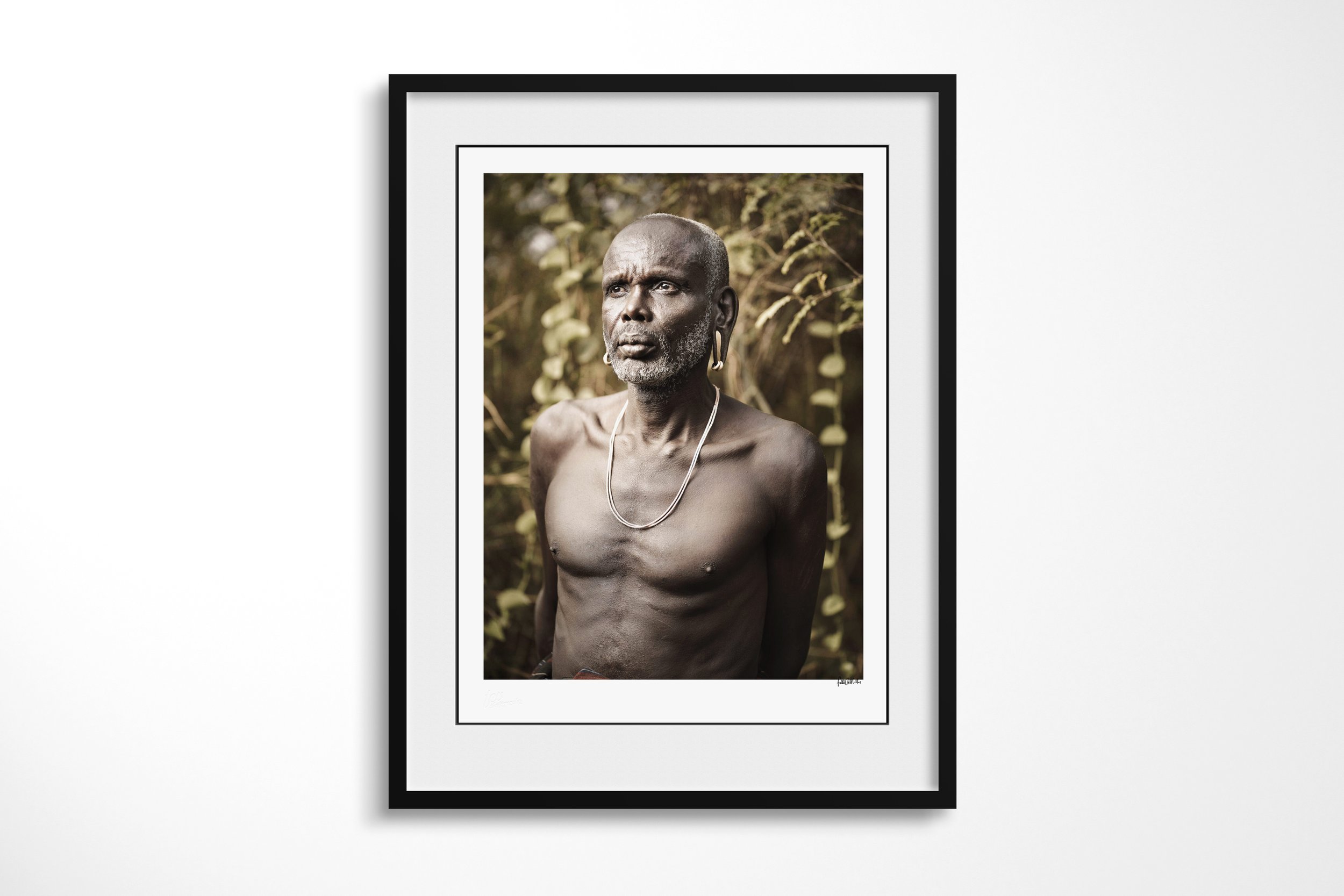 Framed presentation of Bardhoro in a black frame and white mount, showing an older Mursi man shown bare-chested in a close portrait against soft woodland.