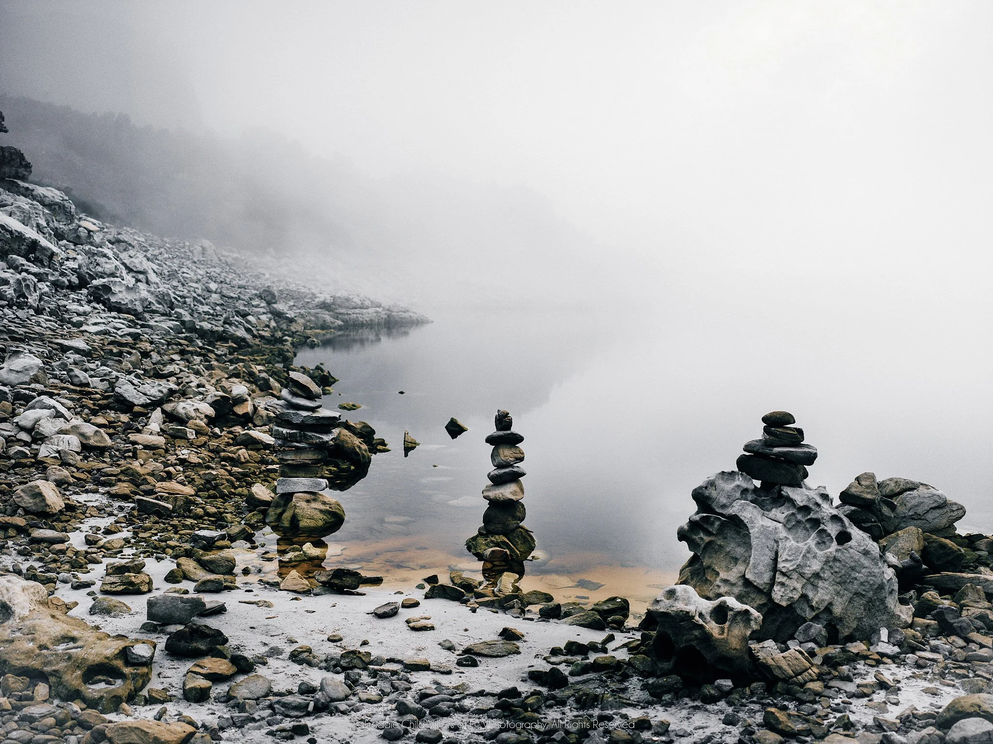 Balanced stone cairns beside still water on a rocky shore fading into pale fog.
