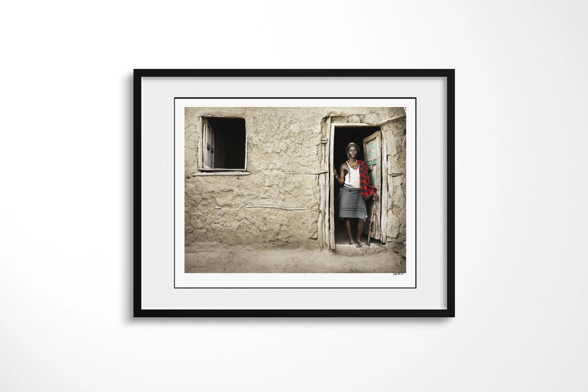Framed presentation of Arboro in a black frame and white mount, showing an Arbore man standing in the doorway of a mud-walled house wearing beadwork and a checked cloth.