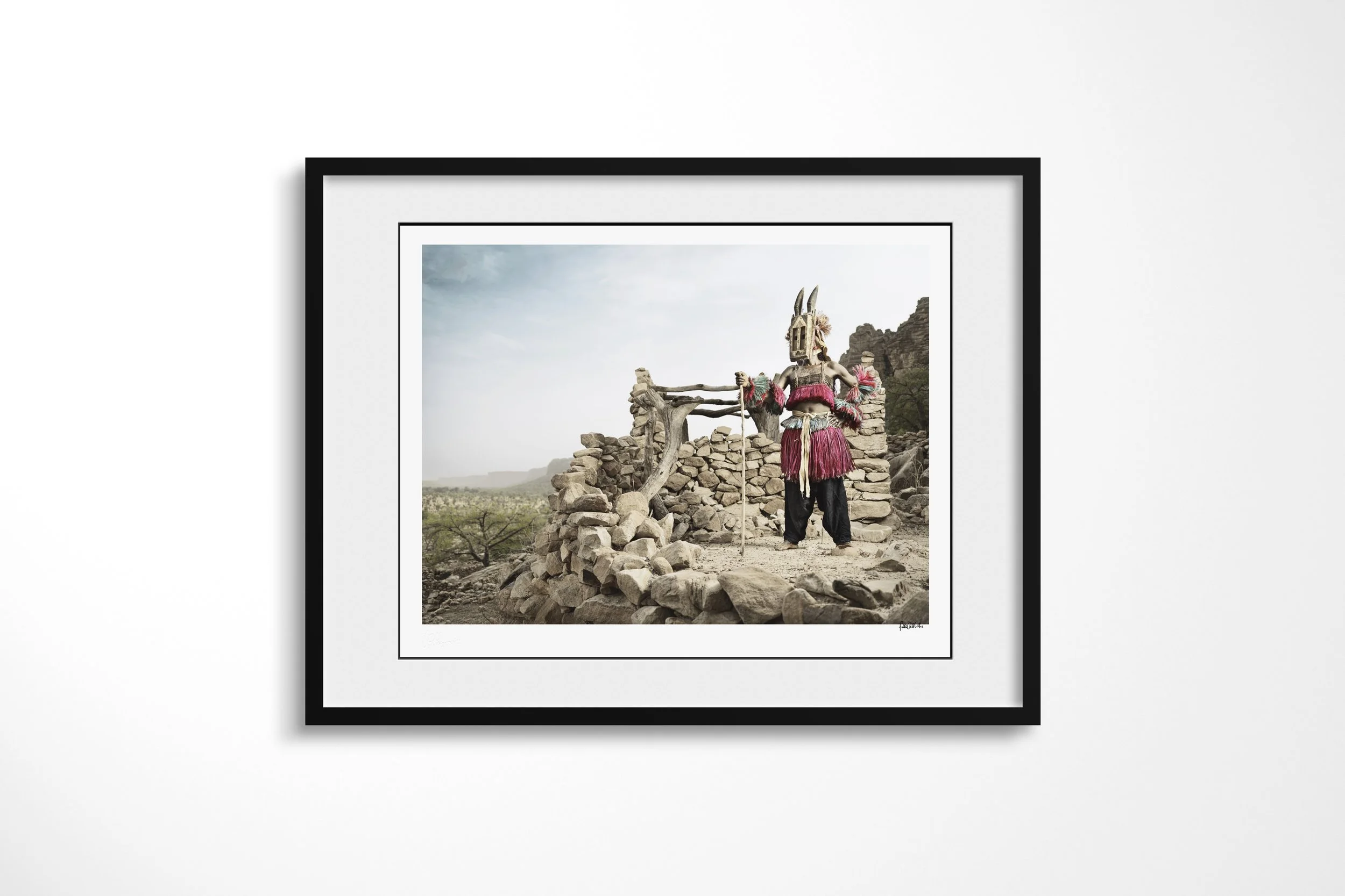 Framed presentation of Walu (Mask) II in a black frame and white mount, showing a Dogon Walu mask holding a staff among the stone walls of a cliff village.
