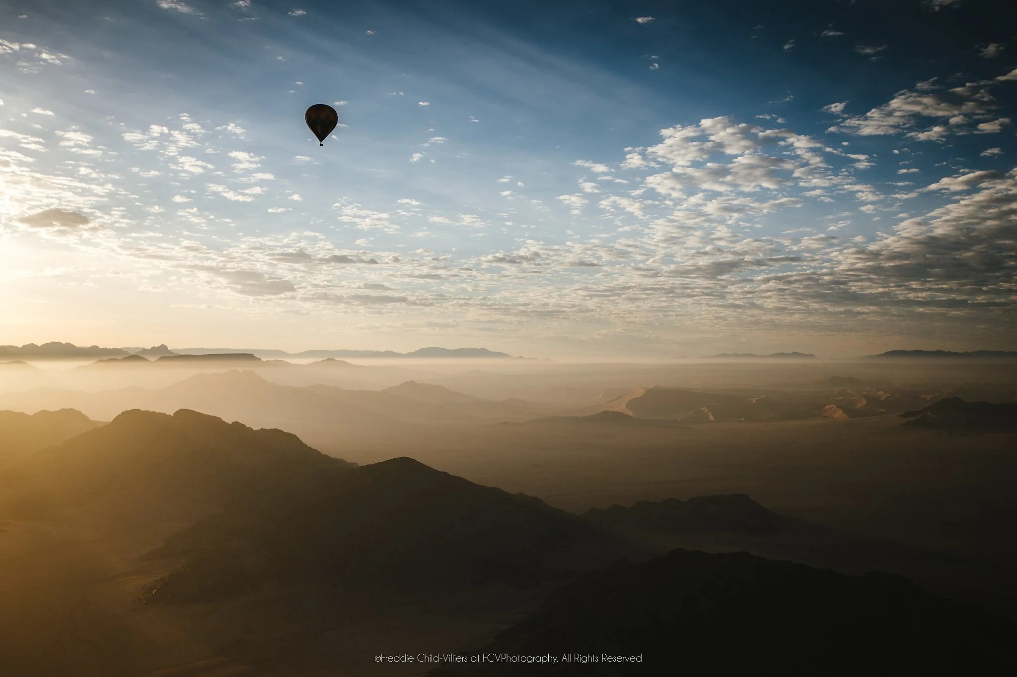 Lone hot-air balloon above layered arid mountain ridges in warm early light beneath scattered clouds.