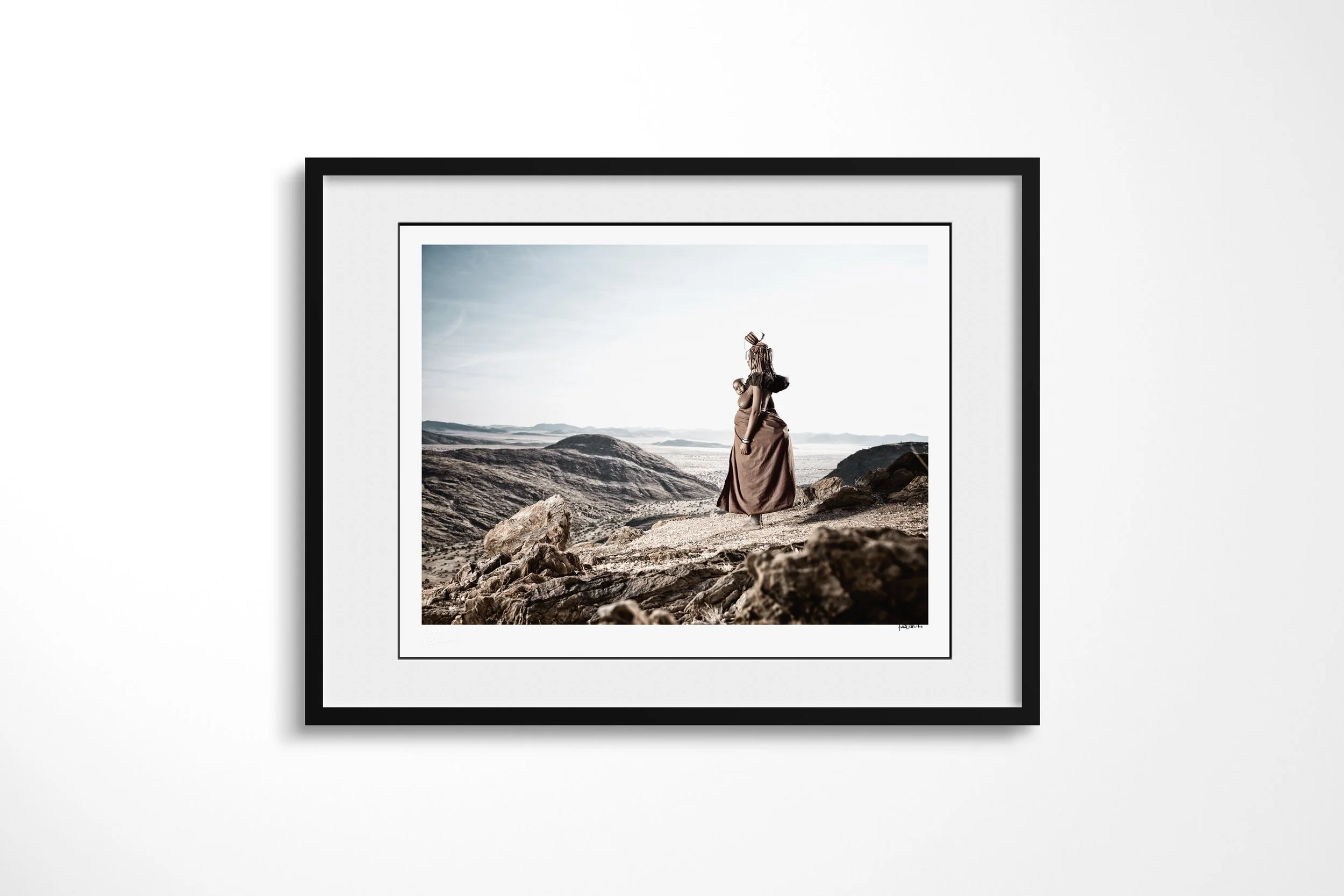 Framed presentation of Venongore in a black frame and white mount, showing Venongore carrying her child on her back while looking across the rocky Marienfluss.