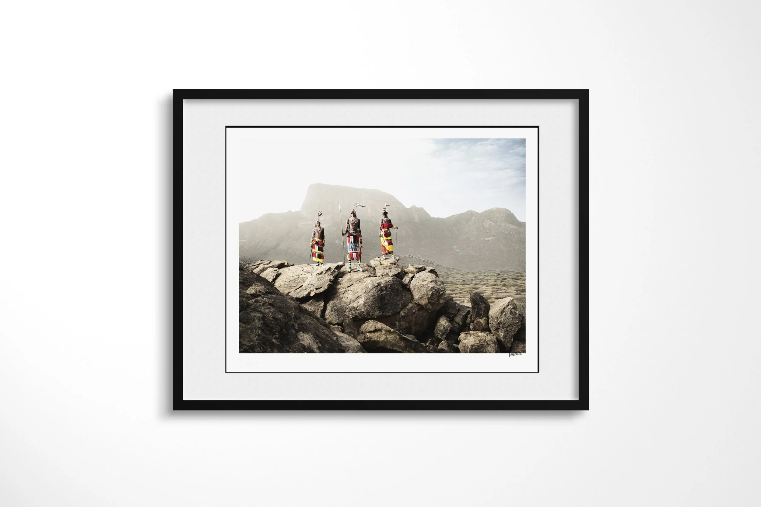 Framed presentation of “The Warriors of Mt.Poi” in a black frame and white mount, showing three Samburu warriors in bright dress and feathered headdresses standing on rocks before Mount Poi.
