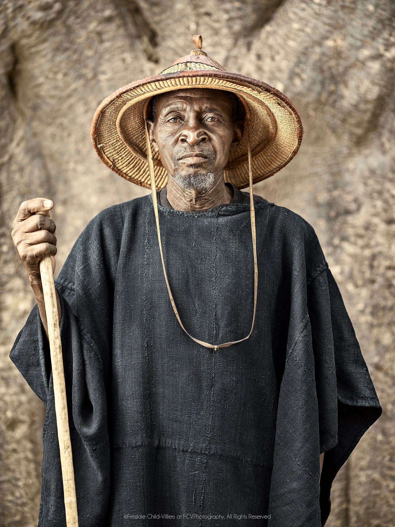 Tigué in a dark robe and broad straw hat holding a staff before a baobab trunk.
