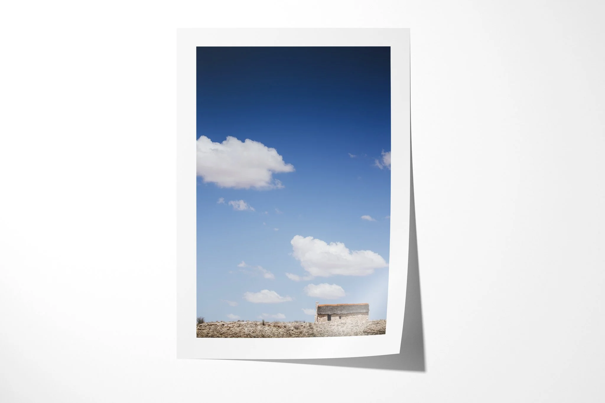 Fine art paper presentation of House On The Hill, depicting a small thatched house isolated on a barren ridge beneath a vast blue sky and scattered white clouds.
