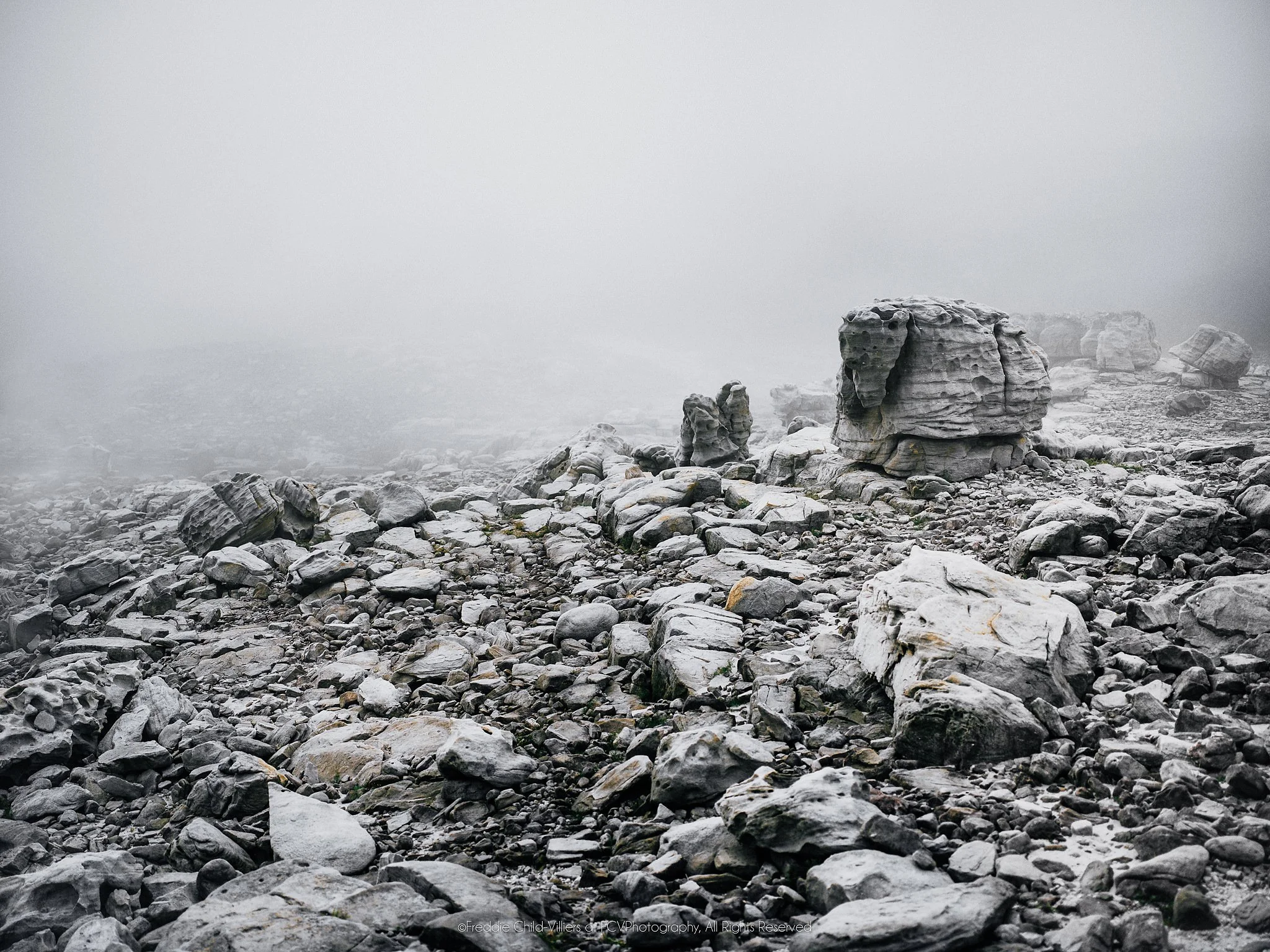 Weathered boulders and broken rock fading into dense mist across a pale mountain landscape.