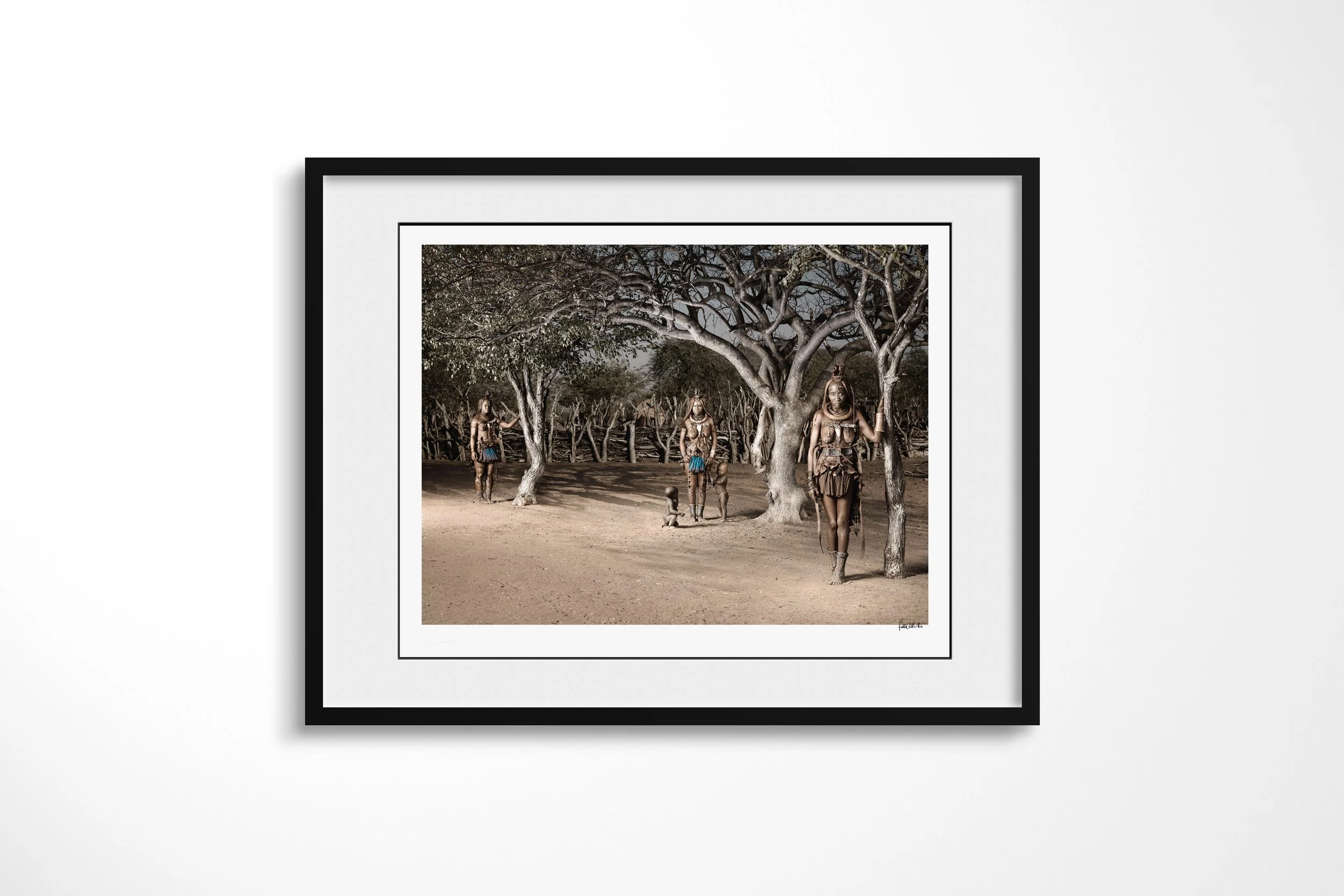 Framed presentation of Waruakawa, Twahenwa and Katitjii in a black frame and white mount, showing three Himba women and children spaced beneath a large tree within their homestead.