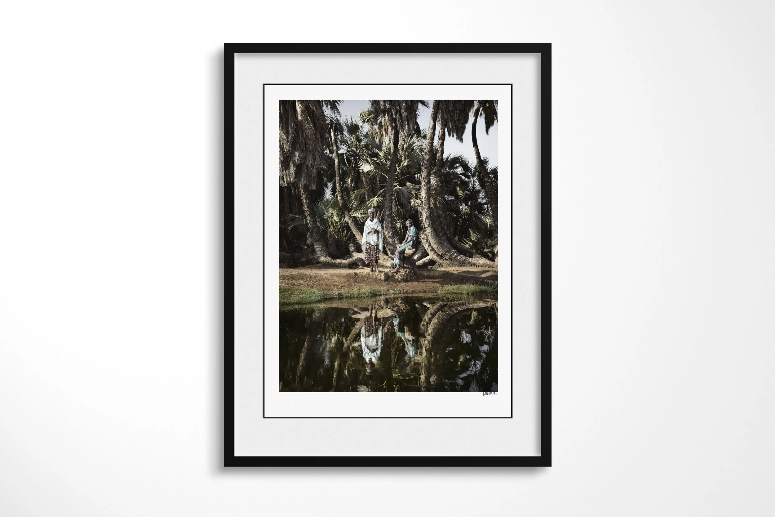 Framed presentation of Talaso & Halkano II in a black frame and white mount, showing Talaso and Halkano standing beside a still spring among dense doum palms.