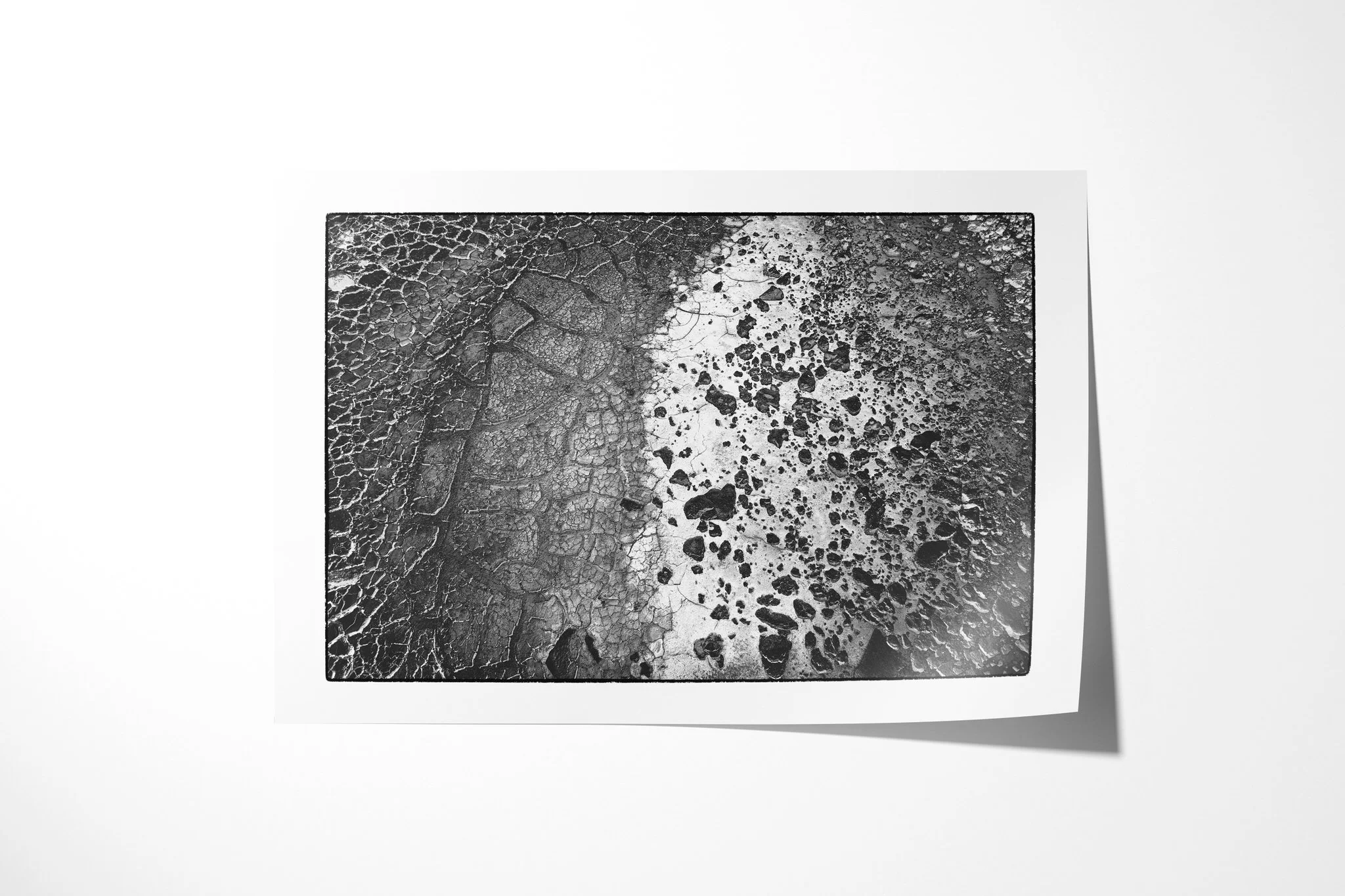 Fine art paper print of Chalbi Desert, showing cracked earth in black and white, divided by a broad pale curving band scattered with dark stones.